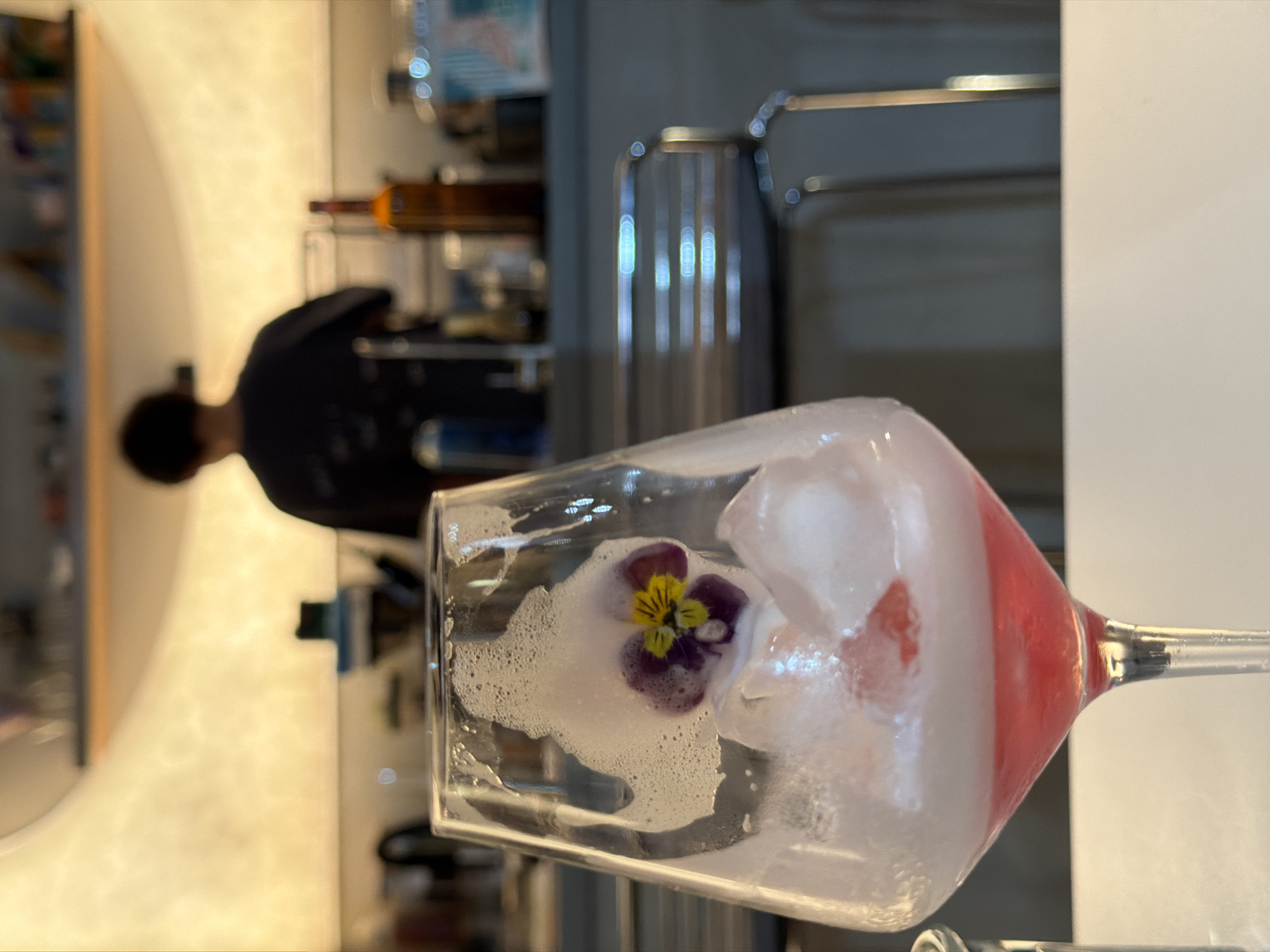A cocktail glass dominates the foreground, containing a white, foamy liquid, ice cubes, a small flower with purple and yellow petals, and a red gradient towards the base of the glass. The glass is slightly tilted. In the background, a blurred figure wearing a black shirt stands in a brightly lit area. Additional blurred background elements include shelves with bottles and possibly a coffee machine. The location is identifiable as Bishkek City, Kyrgyzstan.