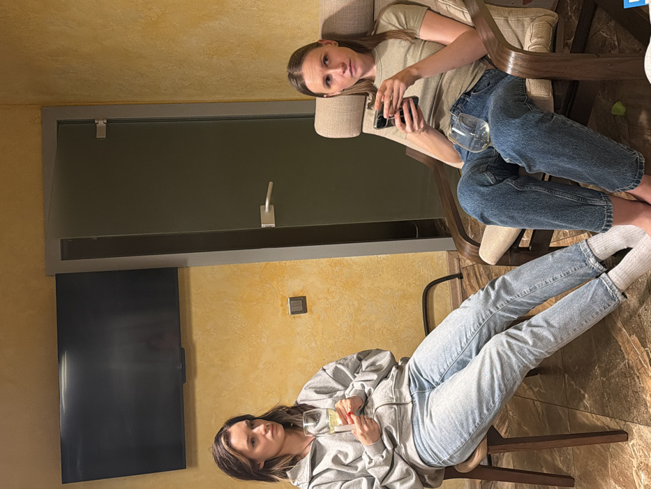 Two women are captured indoors in a casual setting. One woman, seated in an armchair, wears a neutral top and jeans, holding a phone. The other, in jeans and a grey zip-up hoodie, sits on a chair, with a wine glass in hand. A TV is visible, along with a closed door and light switch. The room features yellow walls.