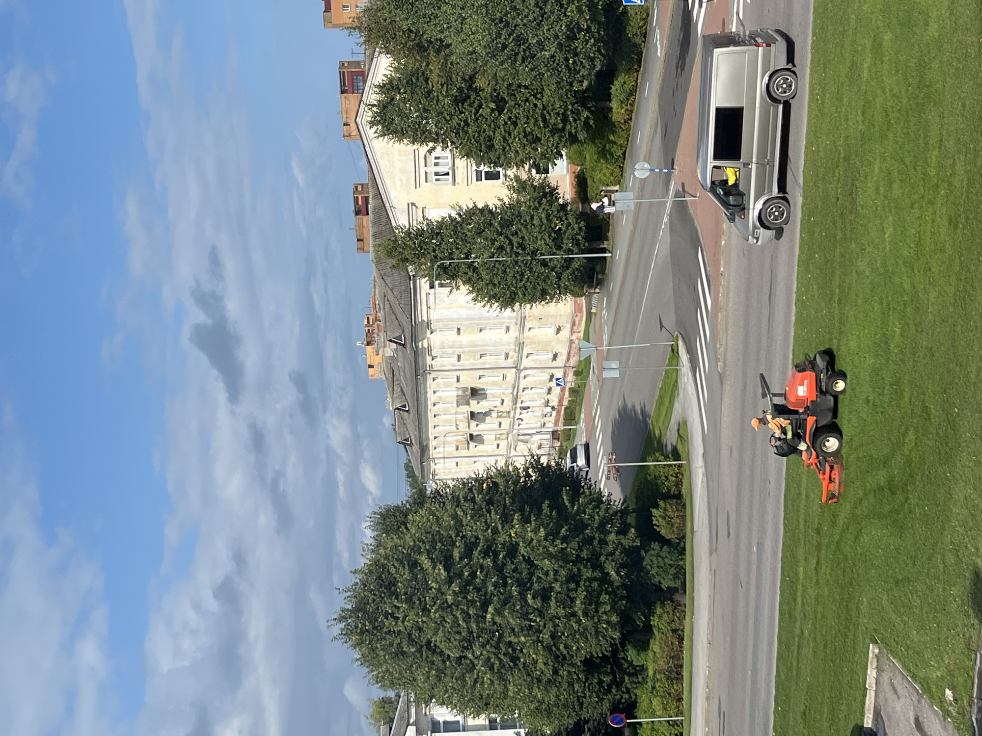 In Kohtla-Järve linn, Estonia, a person wearing an orange high-visibility vest and cap operates a red zero-turn lawnmower on a vibrant green grassy area. Adjacent to the lawn is a paved road where a silver van is traveling. In the background, large deciduous trees with full green foliage stand tall, flanking a pale yellow, multi-story building with numerous windows and a grey tiled roof. The sky above is a bright blue, interspersed with scattered white clouds. The scene is a clear daytime view of a suburban or urban area.