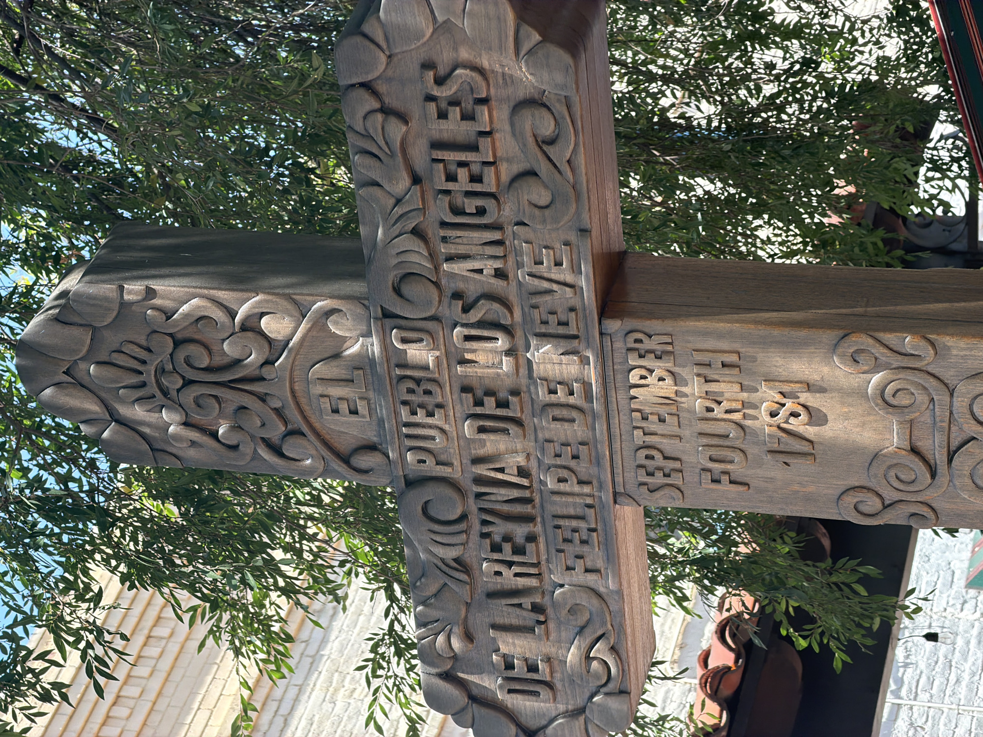 The image shows a close-up of a wooden sign, likely a historical marker, located in El Pueblo de Los Angeles Historical Monument in Los Angeles, United States. The sign is carved with the words "EL PUEBLO DE LA REYNA DE LOS ANGELES FELIPE DE NEVE" on one side and "SEPTEMBER FOURTH 1781" on the other. The carving has a rustic, aged appearance. Trees and glimpses of buildings are visible in the background, indicating an outdoor urban setting. The sign itself is made of dark brown wood.