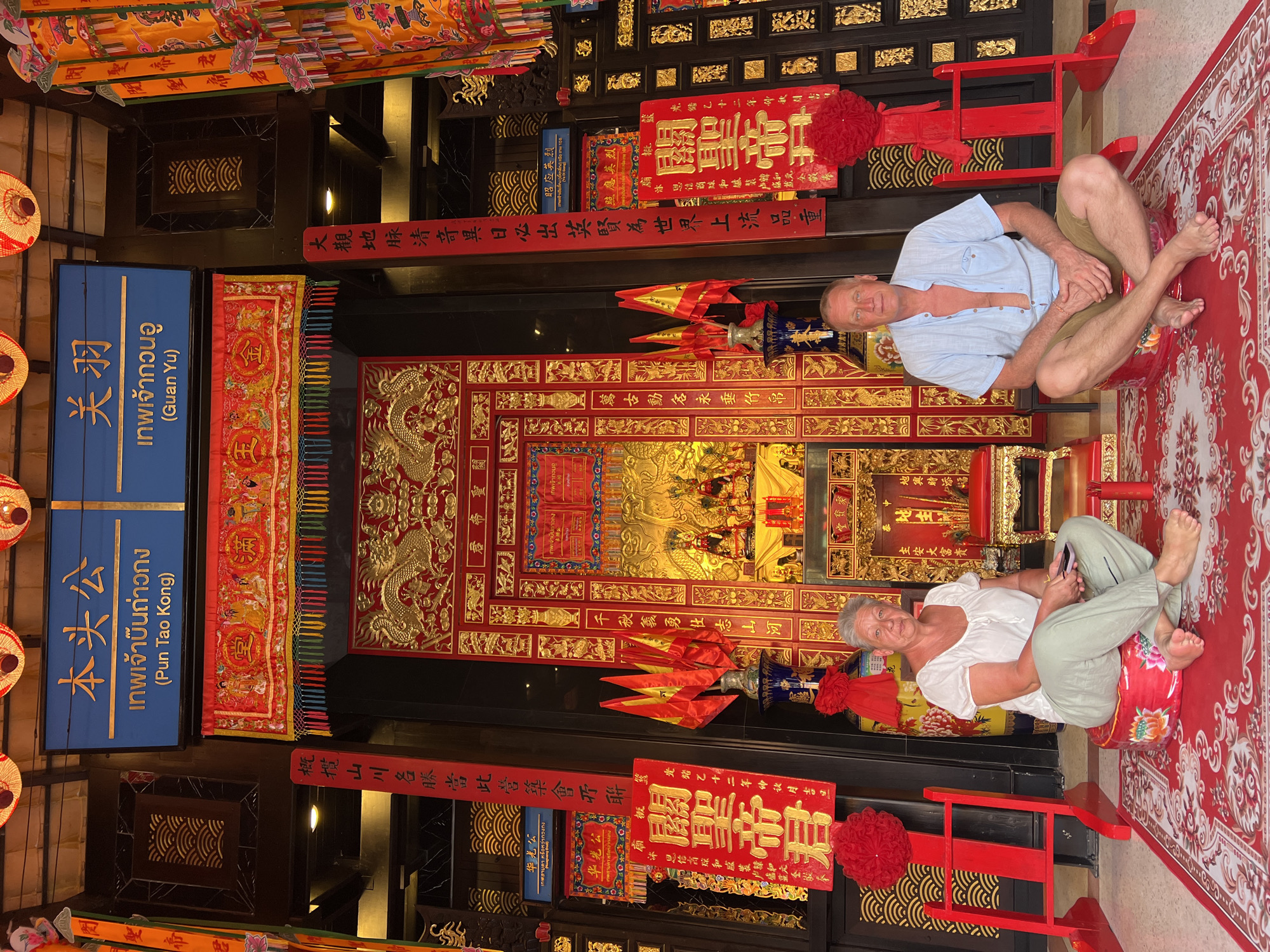 In front of an ornate temple entrance in Baan Hua Thanon, Thailand, a Caucasian couple is seated on red cushions. The woman, on the left, wears a white blouse and light green pants, holding a smartphone. The man, on the right, wears a light blue unbuttoned short-sleeved shirt and tan shorts. They are positioned on a red carpet with white floral patterns. The temple's entrance is elaborately decorated with gold dragons, Chinese calligraphy on red banners, and traditional lanterns. Two red stands with red decorative balls are visible to the sides of the couple. Behind them, the temple interior features a golden altar with statues and religious offerings. The overall scene suggests a visit to a cultural or religious site.