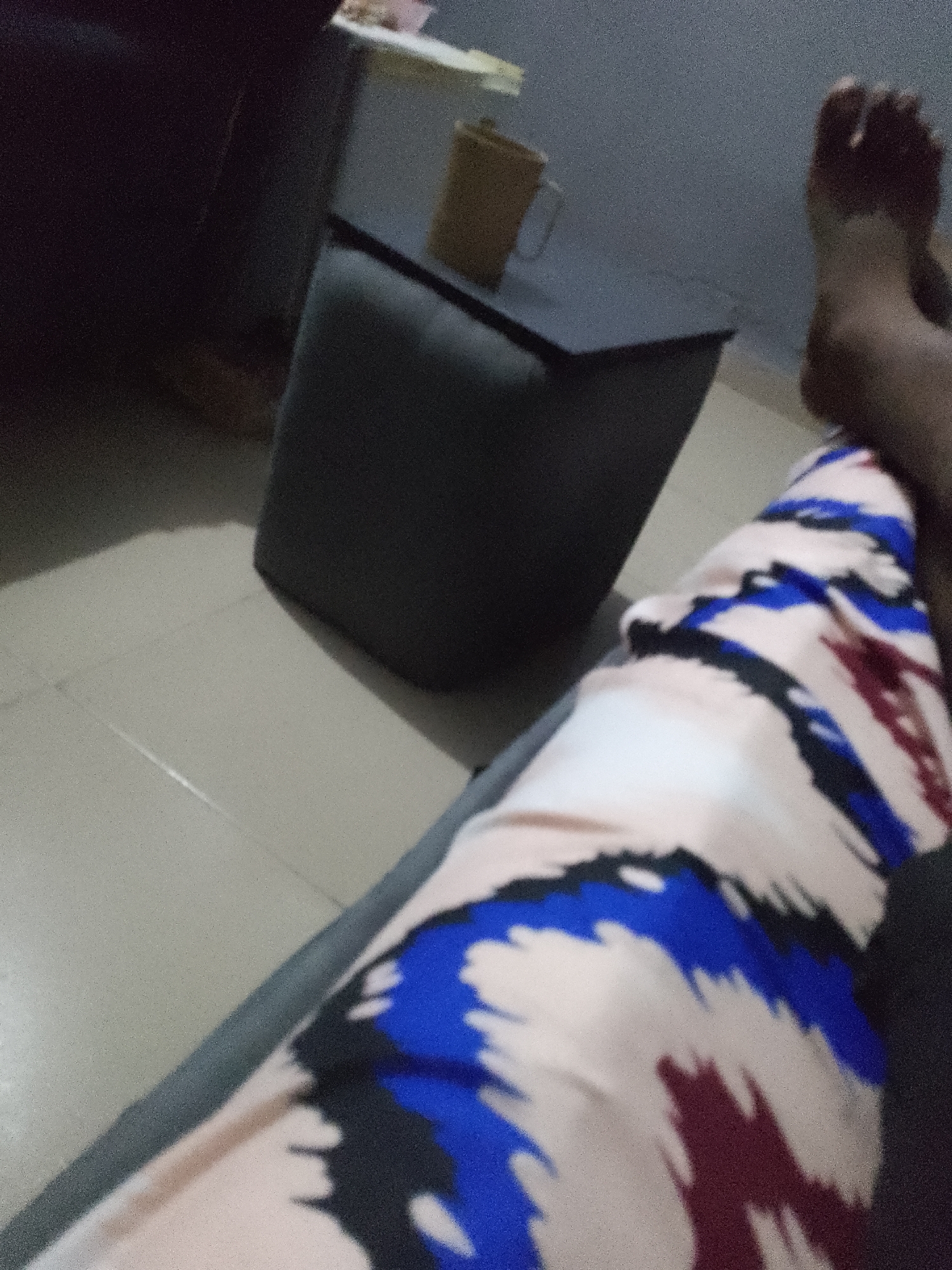 A person's legs are visible, with their feet resting near the right edge of the frame. They are wearing patterned pants with blue, burgundy, and black designs on a light background. A dark gray ottoman or low stool is positioned in the middle ground. In the background, a light-colored cup or mug sits on a dark surface. The floor appears to be tiled. The scene suggests an indoor setting, possibly a living room or lounge area, in Jauro Yinu, Nigeria.