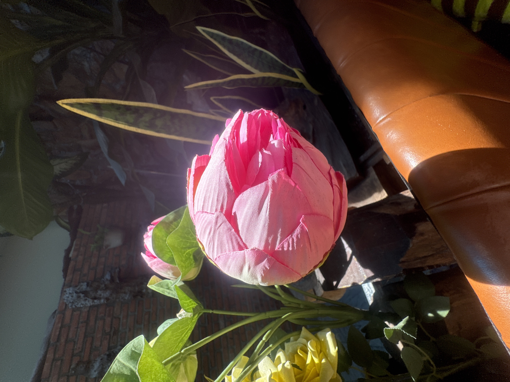 The image shows a close-up of a pink artificial lotus flower bud in the foreground, with its petals tightly closed. To the left of the bud, a bunch of yellow artificial roses are visible, along with green artificial leaves and stems. In the background, a brick wall is partially visible on the left side, and a snake plant with its distinctive striped leaves is seen on the right. Sunlight is casting strong shadows on the scene, particularly on a brown, textured surface that appears to be part of a piece of furniture. The overall setting suggests an outdoor or semi-outdoor decorative arrangement, possibly in a garden or on a patio. There are no people or animals present in the image. The location of Bac Nha Trang Ward, Vietnam, is not directly evident from the visual elements in the photograph.