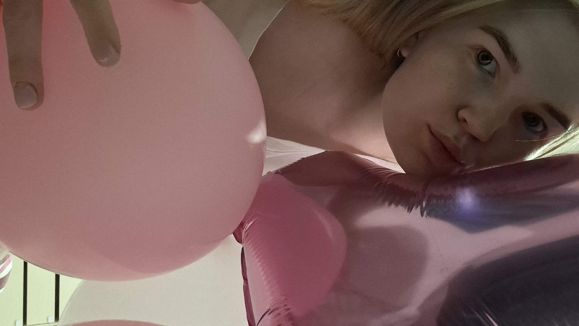 A young, fair-skinned person with light blonde hair is lying down surrounded by pink balloons, seemingly indoors. The person's face, including their eyes, nose, and lips, is visible in the upper right quadrant of the image. One of their hands is holding a large, round pink balloon in the upper left. Another metallic, heart-shaped pink balloon is visible in the lower right quadrant. The background is mostly out of focus, but vertical elements are seen on the left and a white surface is seen in the top-center. The image lacks any details to confirm its location in the city of Chaikovsky, Russia.