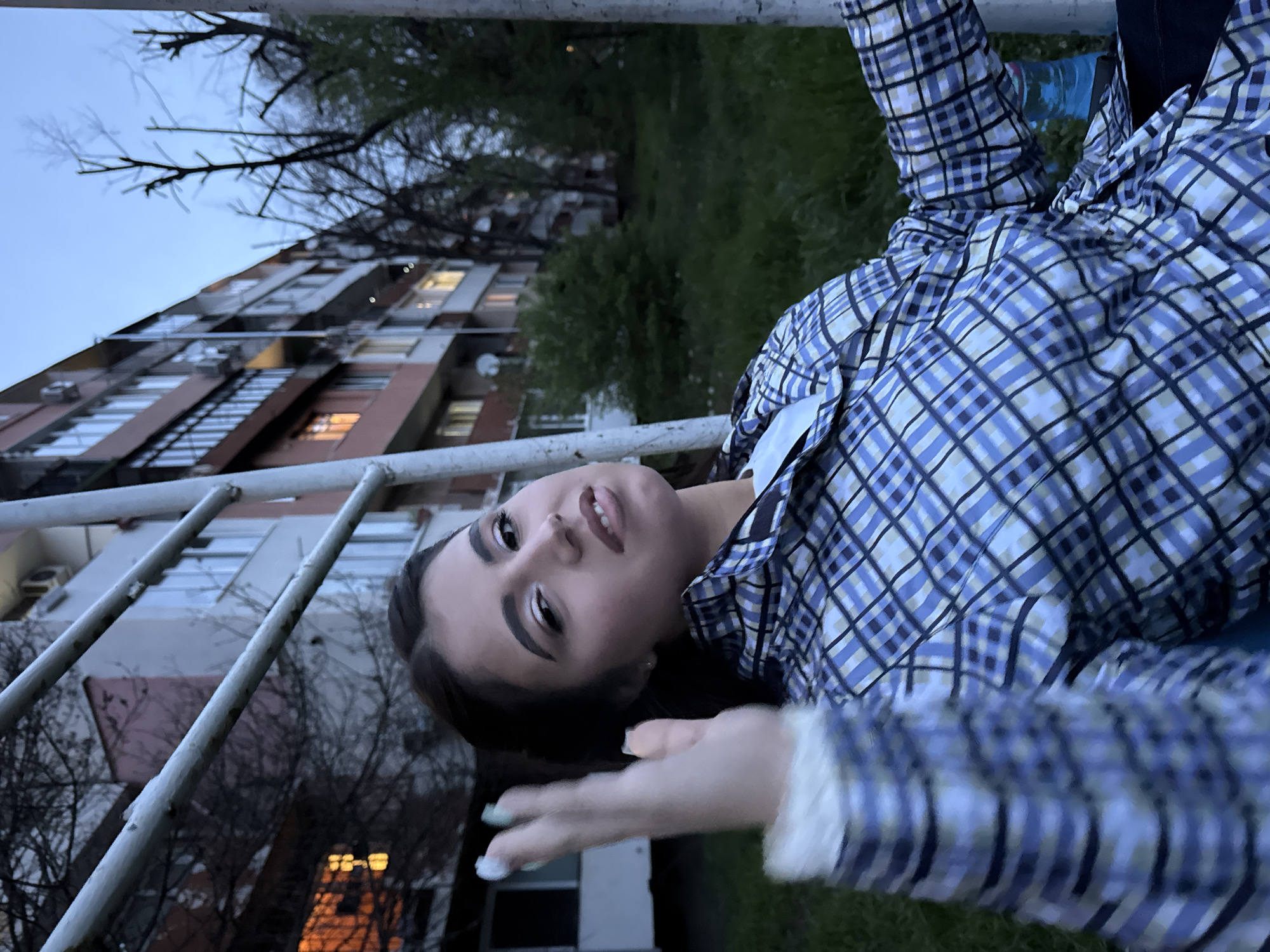 A young woman is lying on a metal structure, possibly playground equipment, in an urban environment. She is wearing a plaid shirt with a white undershirt. Her right arm is outstretched. The background includes a multi-story apartment building with some windows lit, trees, and grass. The photo appears to be taken in the evening or late afternoon. The location is possibly in Sofia, Bulgaria.