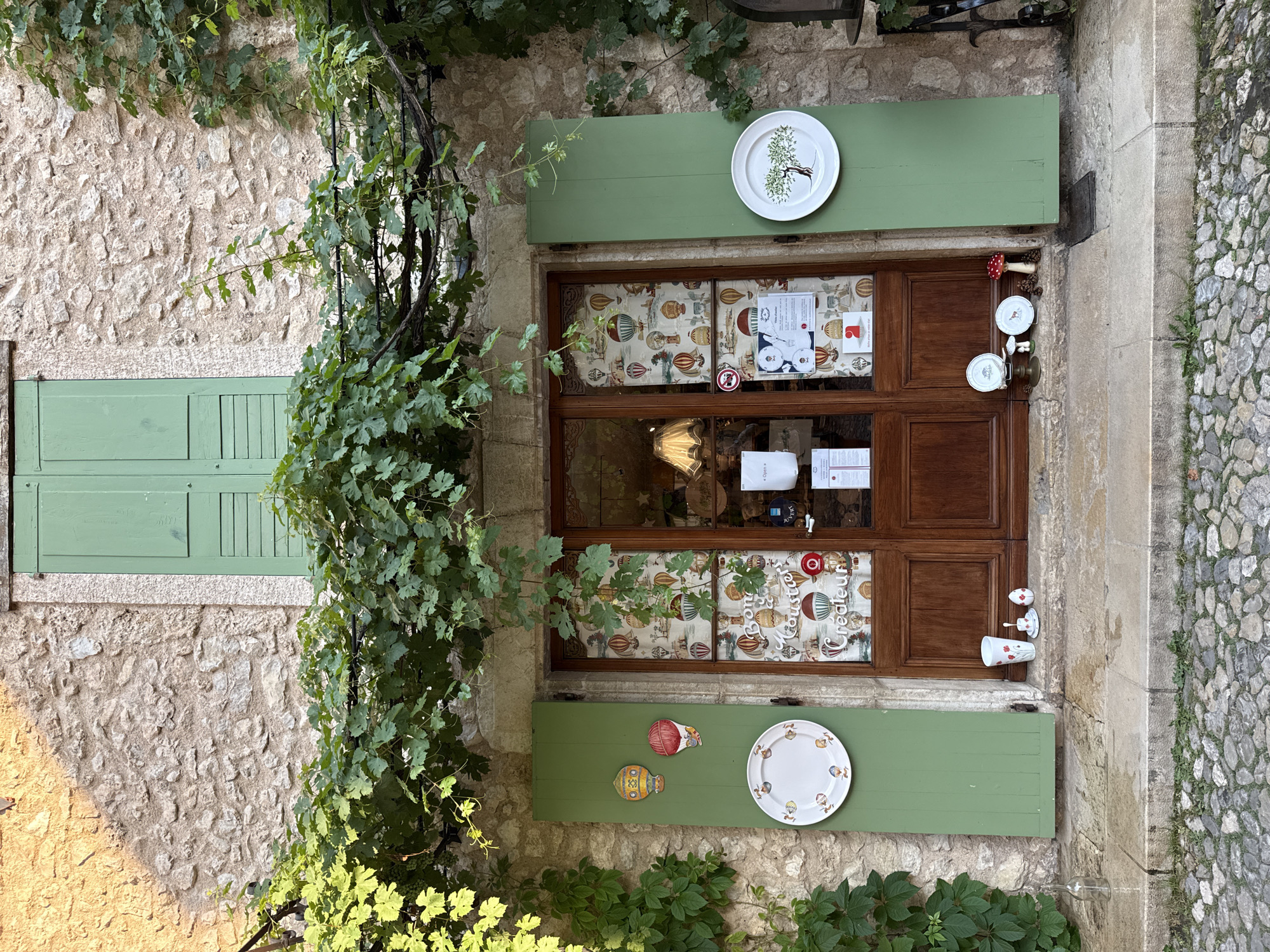 The image shows the exterior of a shop in Moustiers-Sainte-Marie, France. The shop has a wooden double door with glass panes, a stone facade, and green shutters on either side. Above the door, a canopy of green vines hangs down, casting dappled shadows. Decorative plates and hot air balloon motifs are displayed on the shutters and in the shop window. Small decorative items, including cups, saucers, and mushroom figures, are placed on the doorstep. The ground in front of the shop is paved with cobblestones. The lighting suggests it is daytime, with sunlight illuminating parts of the scene. There are no people visible in the image.