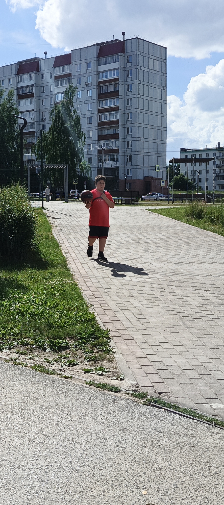 A young person in a red t-shirt and black shorts, holding a basketball, walks on a paved path in front of a tall apartment building. The building has a gray facade with red roof accents and numerous windows. Green trees and grass are visible on the left side of the path, and benches are placed in the background. The sky is blue with scattered white clouds. The scene appears to be in a residential area of Chaikovsky, Russia, likely a park or courtyard.