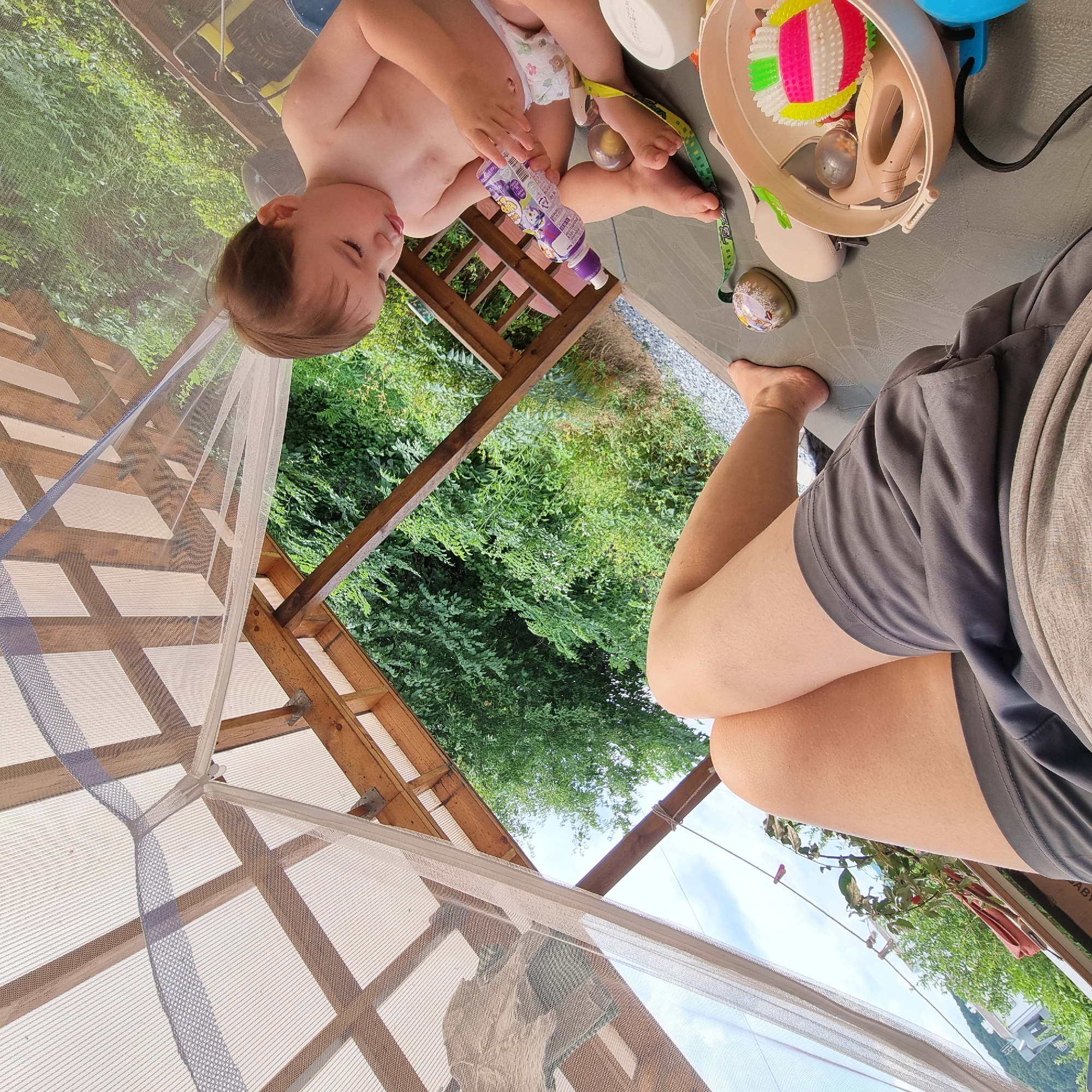 A toddler, wearing a diaper, is playing outdoors on a gray surface. The toddler is holding a purple bottle with a white cap and appears to be looking at it. In the background, there is lush green foliage and a wooden structure, possibly a gazebo or covered patio, with a white mesh covering. A person's legs, wearing gray shorts and a light-colored shirt, are visible in the foreground on the right, suggesting they are sitting or lying down. A blue object, possibly a fan, and several toys, including a colorful spiky ball and a smaller decorative ball, are scattered around the gray surface. The scene appears to be set during the daytime, with natural light illuminating the area. The presence of the wooden structure and greenery suggests an outdoor setting in a residential or recreational area. There is no visible text in the image. The overall mood appears relaxed and casual, with the toddler engaged in play.