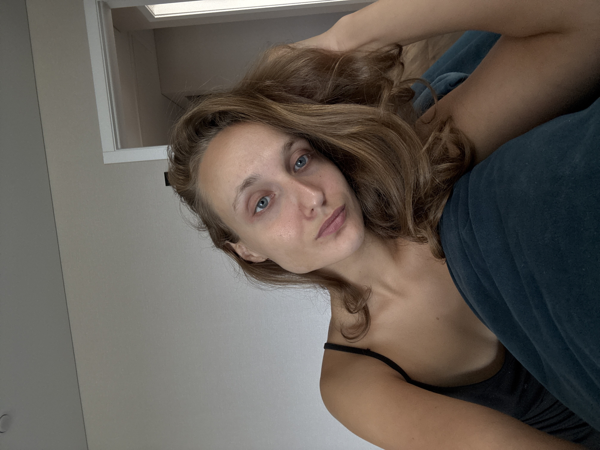 A young woman with light brown hair and blue eyes is photographed from a close-up perspective, looking directly at the camera. She is wearing a black tank top and is partially obscured by a dark teal fabric, possibly a blanket or sofa. Her right hand is raised and resting on her hair near her head. The background is a plain, light-colored wall, with a sliver of a doorway visible on the right side. The lighting suggests an indoor setting, possibly a home. There is no indication of the city of Подшивалово, Russia, in the image.