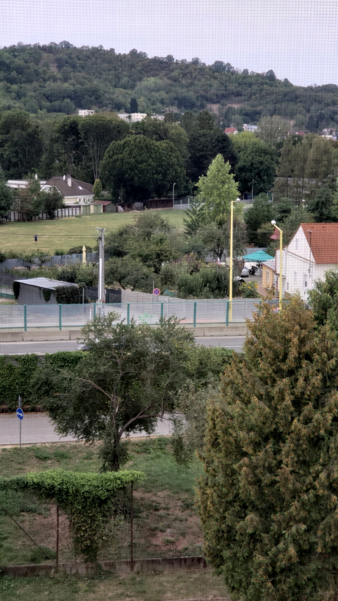 The image depicts a view of a suburban area with a hillside covered in trees in the background. In the middle ground, there is a road separated by a green metal fence from a grassy area with several buildings, including houses and sheds. A tall utility pole with a light fixture stands near some trees. In the foreground, there are more trees and vegetation, with a chain-link fence and a concrete barrier. A blue traffic sign with a white arrow is visible on the left side of the image. The sky is overcast. The location is the Solivarskej Street area in Prešov, Slovakia, as suggested by the presence of the residential buildings, trees, and infrastructure typical of this part of the city. No people are clearly visible in the scene.