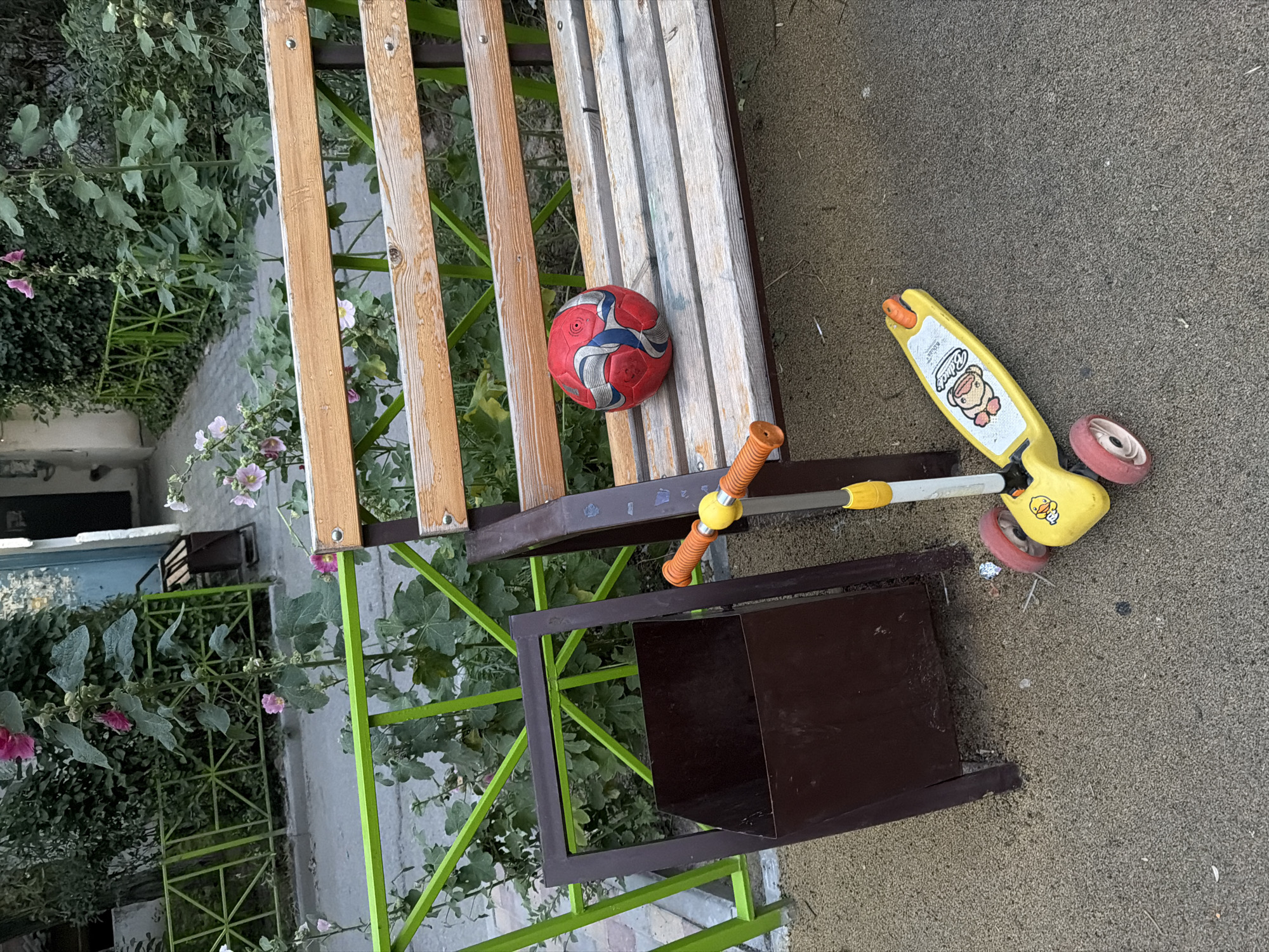 The image depicts a scene in an urban courtyard in Shymkent, Kazakhstan. In the foreground, a yellow child's scooter with three wheels and orange handles is parked on a sandy surface. To the left of the scooter is a dark brown, rectangular trash bin. Behind the scooter and trash bin, a wooden bench with metal supports is situated. A red and blue soccer ball rests on the bench. The background features a green metal fence, and beyond that, lush green foliage and flowering plants, indicative of a well-maintained outdoor space. A building facade is partially visible in the upper right corner.