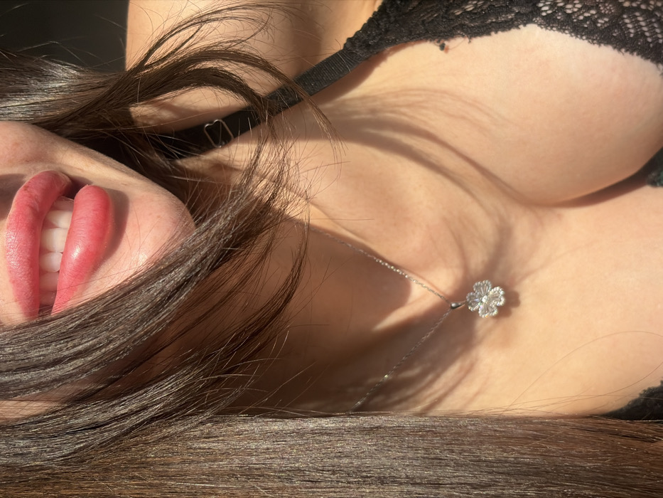 A close-up image features an individual's upper chest and lower facial area. The subject's mouth is slightly open, displaying white upper teeth and full, pink-tinted lips. Long, dark brown hair frames the left side of the face and extends over the shoulders. A silver-colored chain necklace with a four-leaf clover pendant, embellished with clear, reflective stones, rests on the visible decolletage. A black lace bra and a thin black bra strap are visible on the right side of the frame. Strong directional light illuminates the subject, casting shadows across the neck and chest. The background is uniformly dark and out of focus. The image provides no visual evidence to confirm the specified location of Saint Petersburg, Russia.
