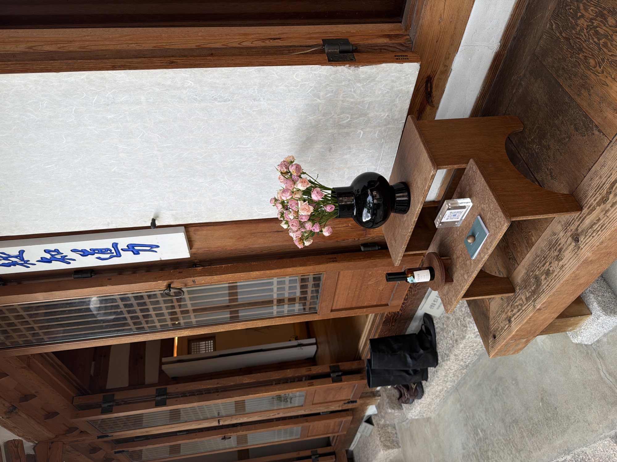 A photograph captures the exterior of a traditional Korean building in Seoul, South Korea, likely a hanok. The building features wooden doors with latticed glass panels, which are open, revealing a glimpse of the interior. A white paper screen is visible on the right side of the building. In front of the building, a multi-tiered wooden display stand holds a black vase with pink roses, a small bottle of what appears to be wine, a small transparent case with a QR code, and a small green book or notebook. Boots and shoes are placed on a stone step to the left of the display. The overall scene evokes a sense of traditional Korean aesthetics and hospitality.