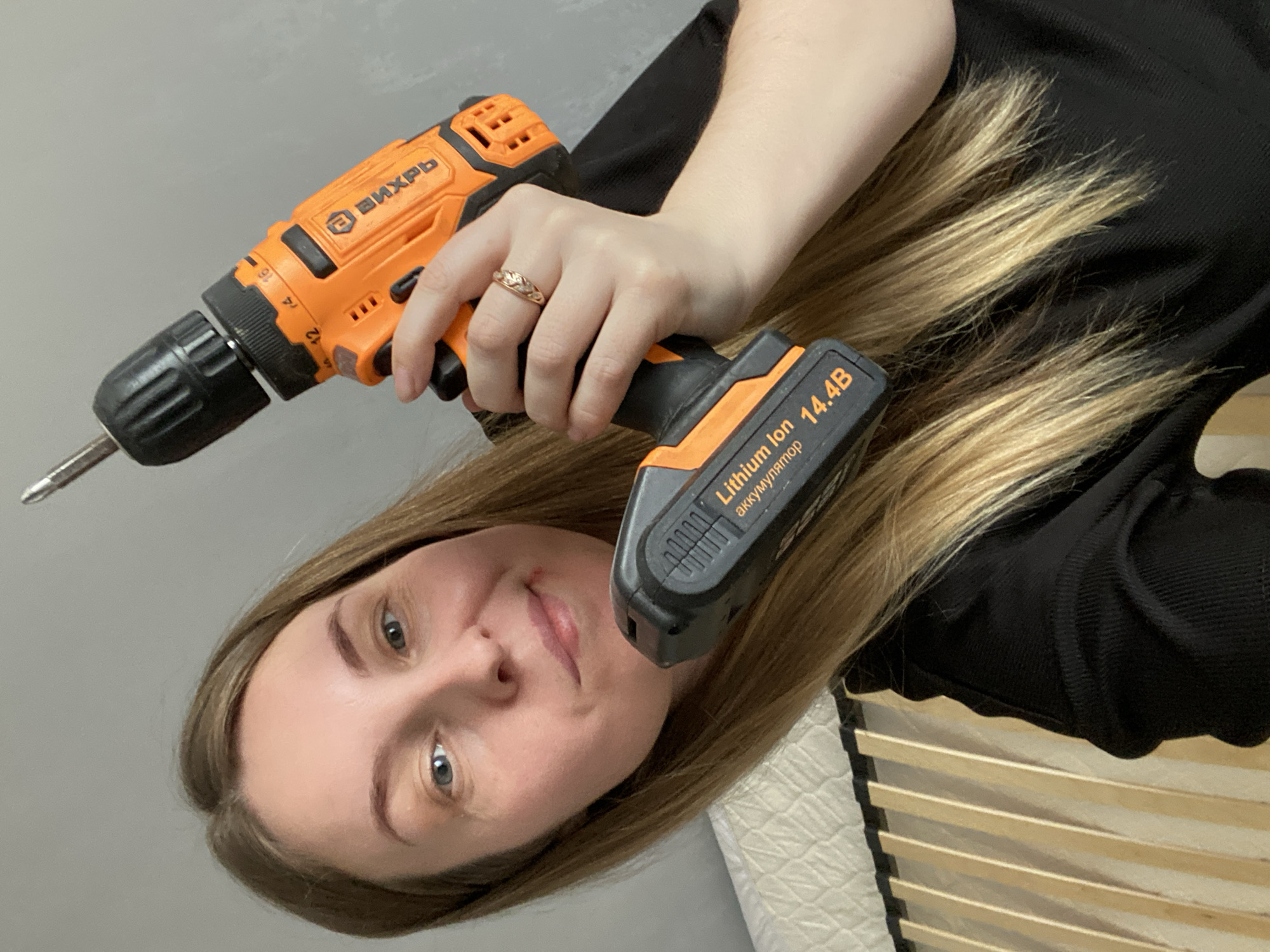 A woman with blonde hair is holding an orange and black power drill. The drill has a battery pack labeled "Lithium Ion 14.4B". The woman is wearing a black shirt and a ring on her finger. The background is a gray wall and a wooden bed frame. The image was likely taken indoors. There is no information suggesting this image was taken in Noviy, Russia.