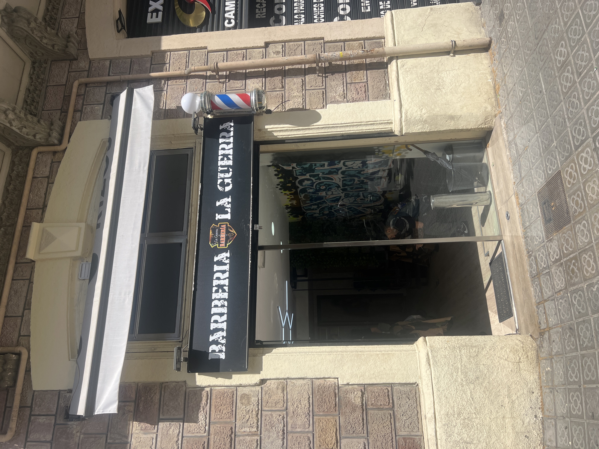 The image captures the entrance of a barber shop named "Barberia La Guerra" in what appears to be the city of Barcelona, Spain, as indicated by the distinctive flower-patterned paving stones on the sidewalk, typical of Barcelona's urban design. The barber shop features a traditional red, white, and blue barber pole mounted above a black sign that vertically displays the shop's name in white lettering, "BARBERIA LA GUERRA". A small logo is visible on the sign. The shop has a glass door and a window with a white awning. The building exterior is constructed with stone brickwork, and the entrance is framed by decorative stone molding. The image suggests a sunny day and a typical urban setting in Barcelona.