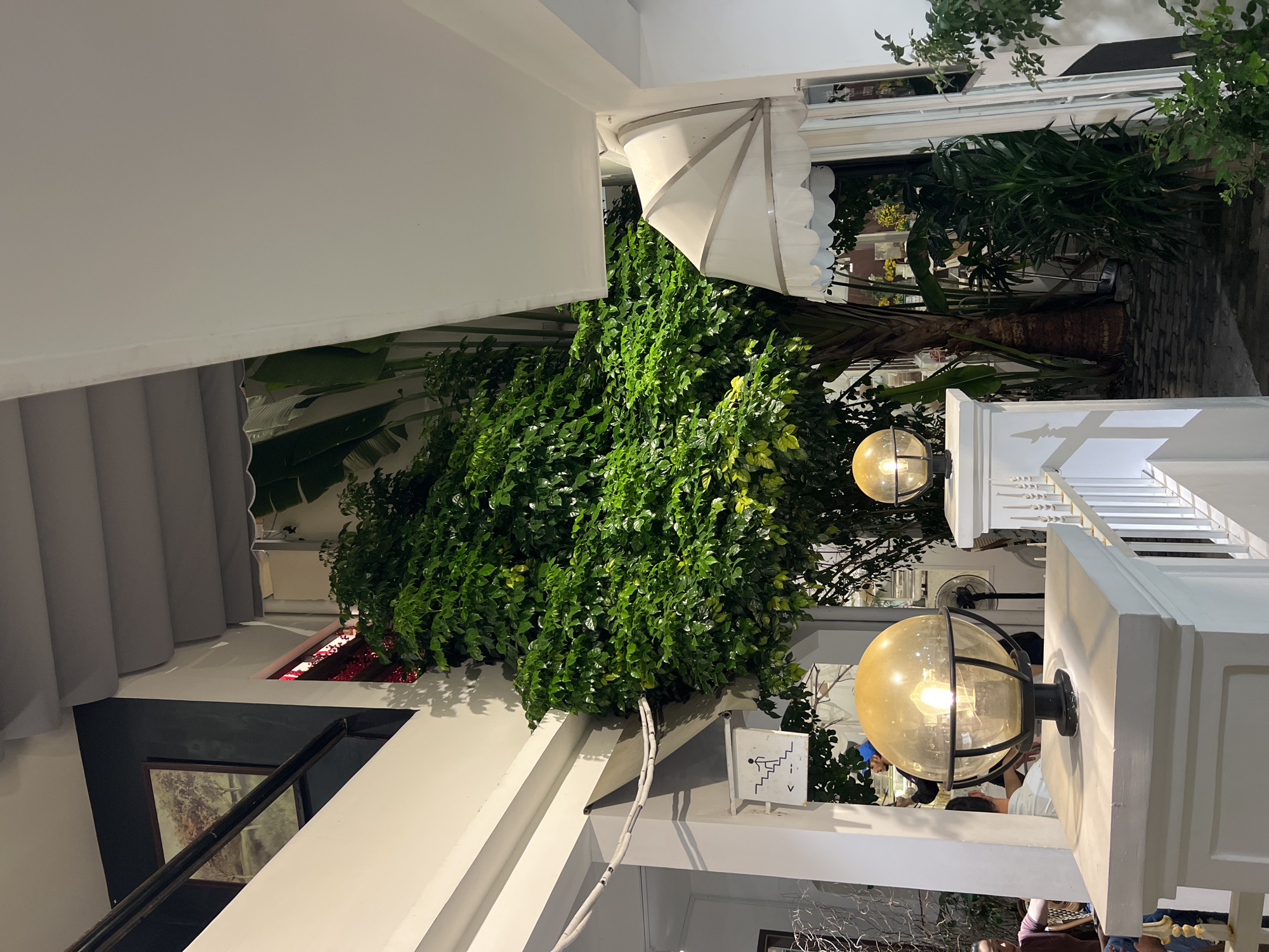 The image shows an indoor/outdoor space, possibly a restaurant or hotel in Nha Trang, Vietnam. The setting features a staircase on the left, next to a painting or artwork displayed on the wall. Overhead, a dense collection of green plants and foliage spills over the edges of what appears to be an open planter box. An awning or architectural detail is visible in the upper-right, adjacent to more greenery. Two spherical light fixtures with metal cages are affixed to the white structural supports on the right side of the image. There is a sign indicating the staircase. People are partially visible in the background.