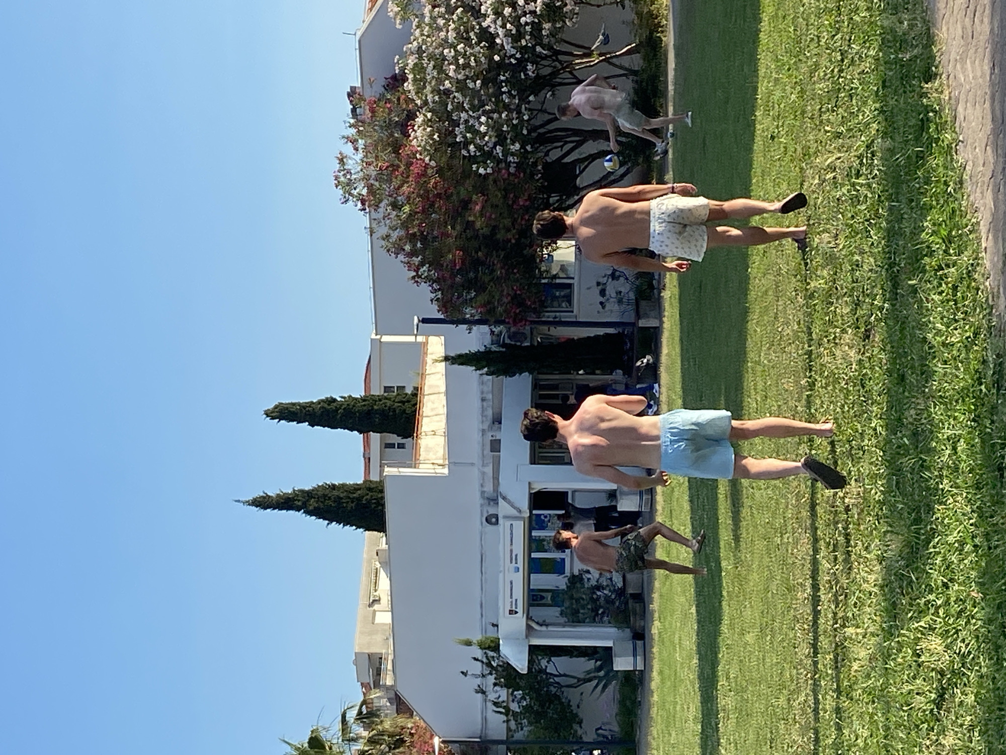 In Budva, Montenegro, two young men are captured in a candid moment on a sunny day, enjoying the outdoors. They are on a well-manicured grassy area, with a paved walkway to the right of the frame. In the background, a modern white building with blue accents stands under a clear, bright blue sky. To the left of the building, lush green trees, including two tall, slender cypress trees, add to the picturesque scenery. A vibrant flowering tree, with a mix of white and pink blossoms, adds a splash of color to the scene.
One man, wearing patterned shorts, is captured from behind, his back to the camera. He appears to be walking on the grass, looking towards something out of frame. Another man, in light blue shorts, is seen from the side, also walking on the grass. His posture suggests he is engaged in a casual activity. Further back, another individual, partially visible, seems to be kicking a volleyball, implying a game is in progress. The overall atmosphere is relaxed and recreational, fitting for a warm, sunny day. The lighting suggests it is daytime, likely late morning or afternoon, given the clear sky and the shadows cast on the grass. The clear sky and vibrant greenery indicate pleasant weather. No visible text is discernible in the image.