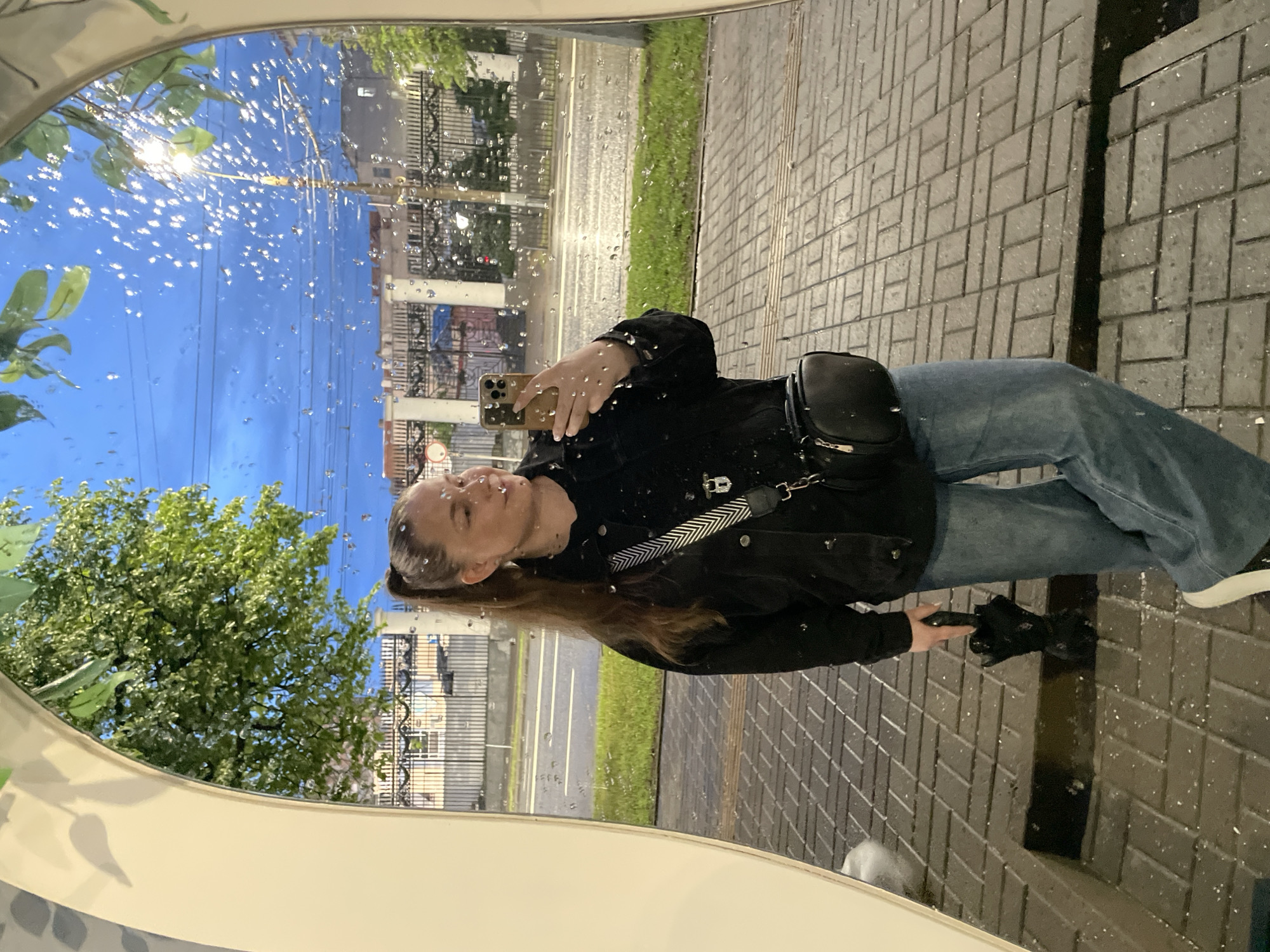 A woman is reflected in a rain-covered mirror, apparently taking a selfie with her phone. She wears jeans, a black jacket, and a crossbody bag. She holds a black umbrella in her left hand. The background shows an urban scene with buildings, trees, a street, and a sidewalk. Based on the reflection in the mirror, the scene includes elements that correspond to the city of Izhevsk, Russia.