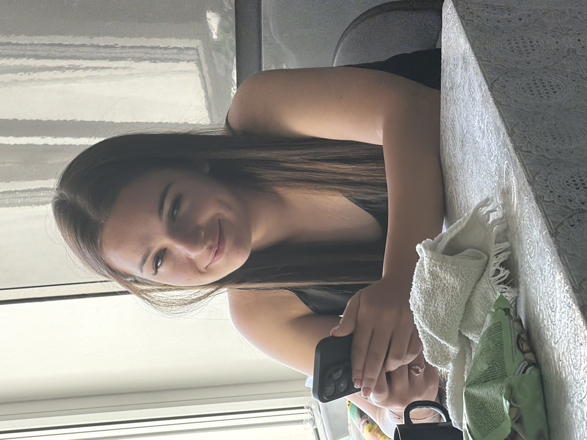 A young woman with long, brown hair and light skin is seated at a table, holding a black smartphone in her hands. She is looking directly at the camera with a slight smile. She is wearing a black, sleeveless top. On the table in front of her are a white towel, a package of what appears to be nuts or snacks, and a black mug. The table is covered with a white lace tablecloth. Behind the woman, a window with vertical blinds is visible, letting in natural light. The background also shows a hint of a chair and a wall. The setting suggests an indoor environment, possibly a home, in the city of Strășeni, Moldova.