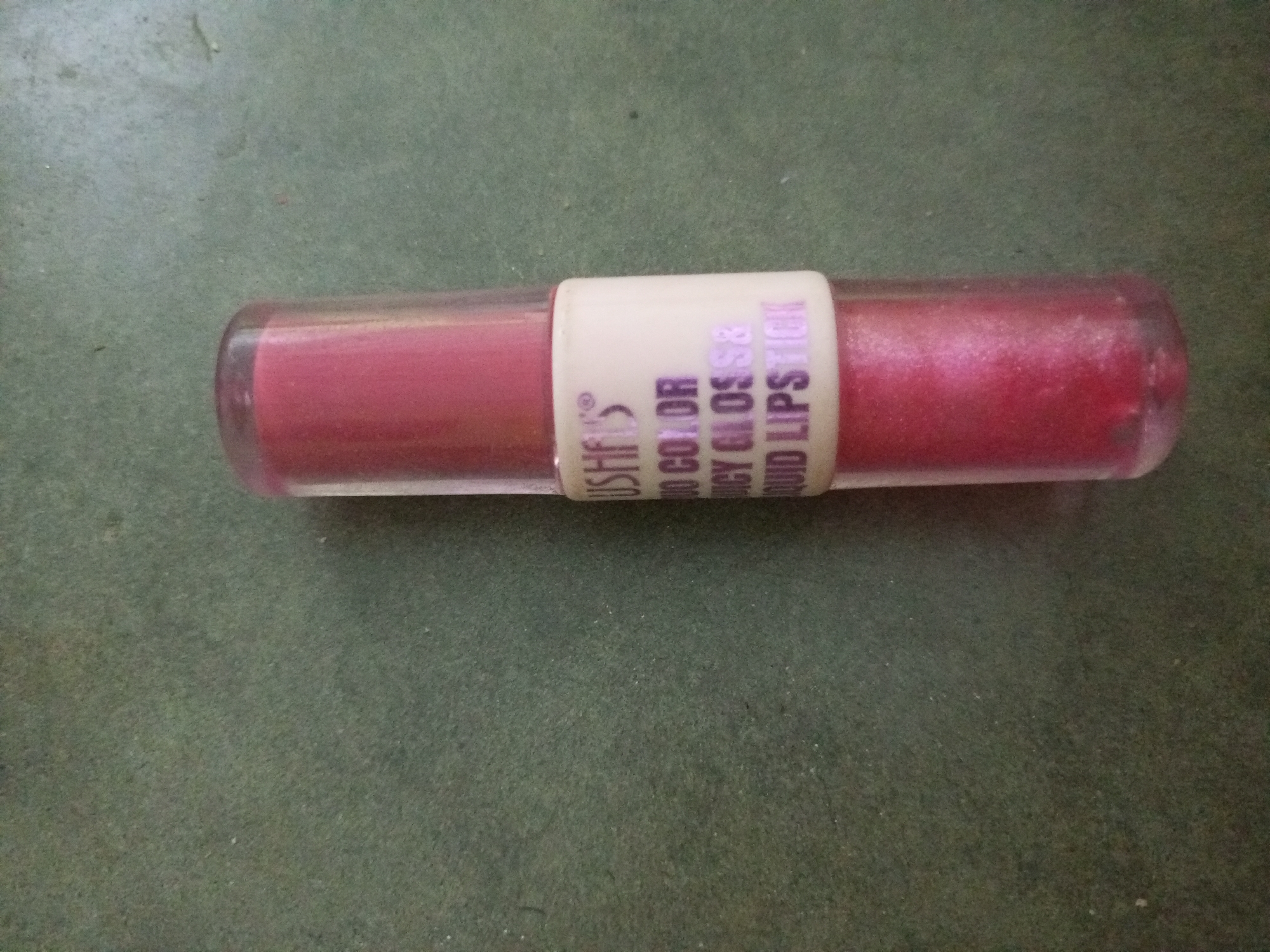 A tube of pink, shimmery liquid lipstick lies horizontally on a green, textured surface. The tube is transparent, revealing the product inside, and has a white label with purple text that reads "USHES 3D COLOR JUICY GLOSS LIQUID LIPSTICK." The scene is indoors, as indicated by the artificial lighting and the consistent background texture. There are no people visible in the image, nor any indication of the specific location within La Tagua, Venezuela. The lipstick appears to be the sole subject of the photograph.
