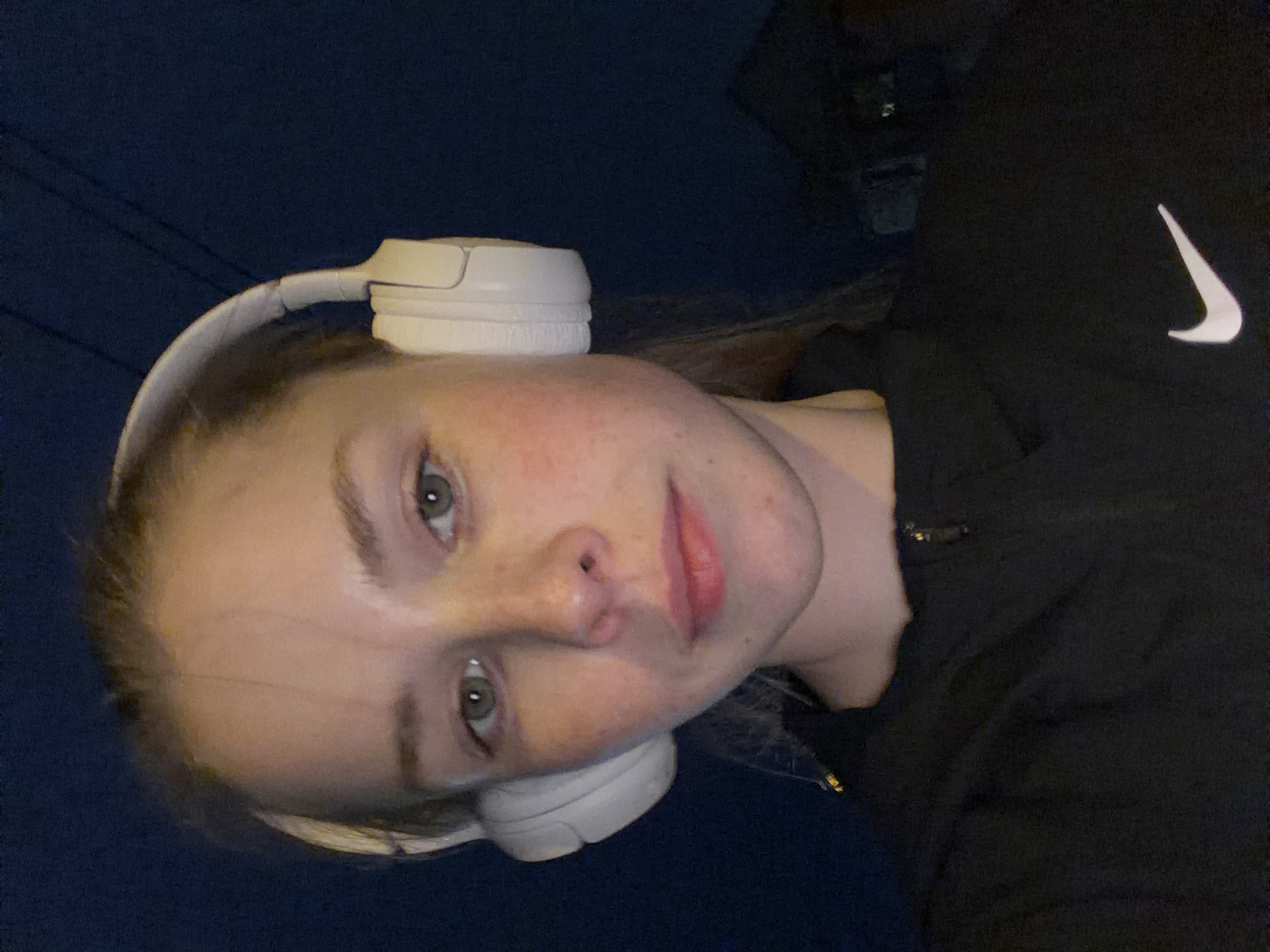 A young woman with fair skin and light brown hair wears white headphones. She has visible freckles and is wearing a black Nike jacket. The background appears to be a dark blue night sky. The photo's location is not identifiable as Berezniki, Russia based on the given image.