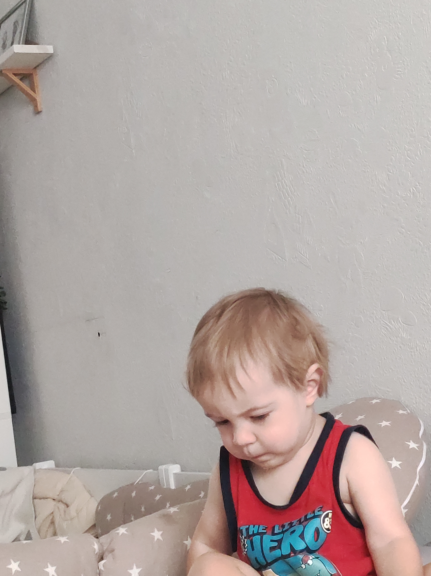 A toddler with blond hair is sitting in a crib. The child wears a red tank top with black trim and a graphic that says "The Little Hero." The crib has tan and white star-patterned bumpers and off-white bedding. The room has light gray walls with a shelf in the top left corner. The location is assumed to be in a domestic environment. The city of Chaikovsky, Russia, cannot be confirmed.