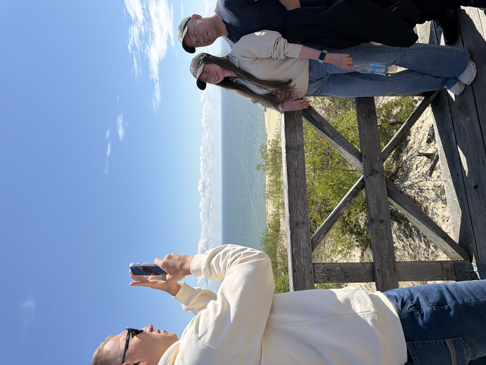 In the city of Morskoye, Russia, three people are on a wooden boardwalk overlooking a beach and the sea. A man in the foreground on the left is wearing sunglasses and a cream-colored hoodie and is holding up a smartphone, appearing to take a picture. To the right, a woman wearing a beige jacket, blue jeans, and a dark baseball cap stands next to a man in a dark t-shirt and a camouflage baseball cap. The woman is holding a bottle of water in her right hand. The sea is calm and a light blue, with a sandy beach in the distance. The sky is clear and blue with a few scattered white clouds. The boardwalk is made of weathered wood and has a railing with a geometric design. Green foliage is visible on the sand dunes.