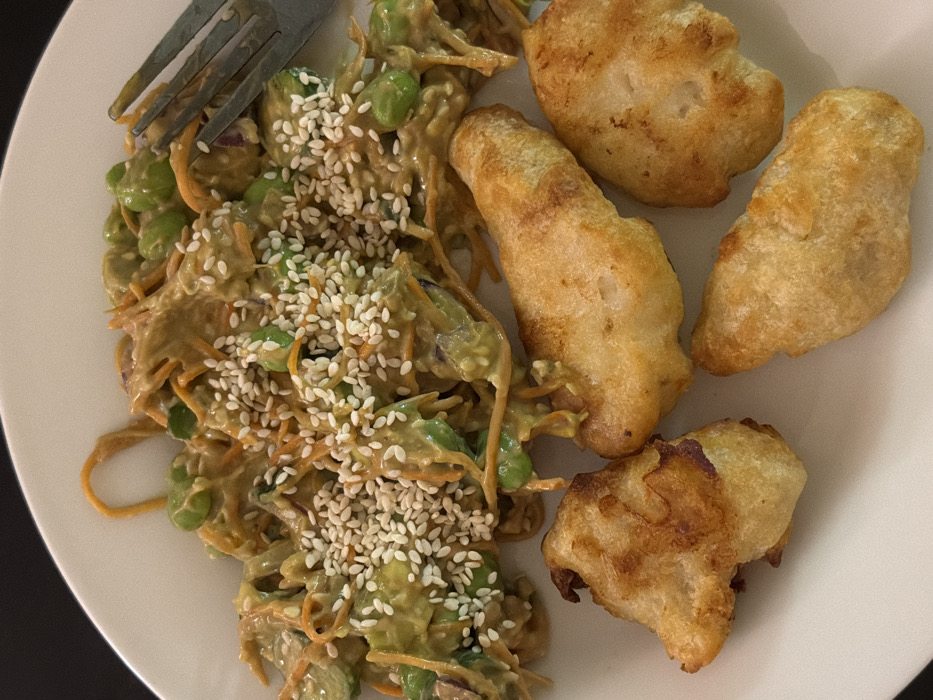 A plate presents a culinary arrangement, featuring a salad dressed with a creamy sauce and sprinkled with sesame seeds, accompanied by a fork. Adjacent to the salad, four pieces of fried food, possibly chicken or fish, rest on the plate, displaying a golden-brown hue. The composition suggests a meal combining a fresh salad with fried components.