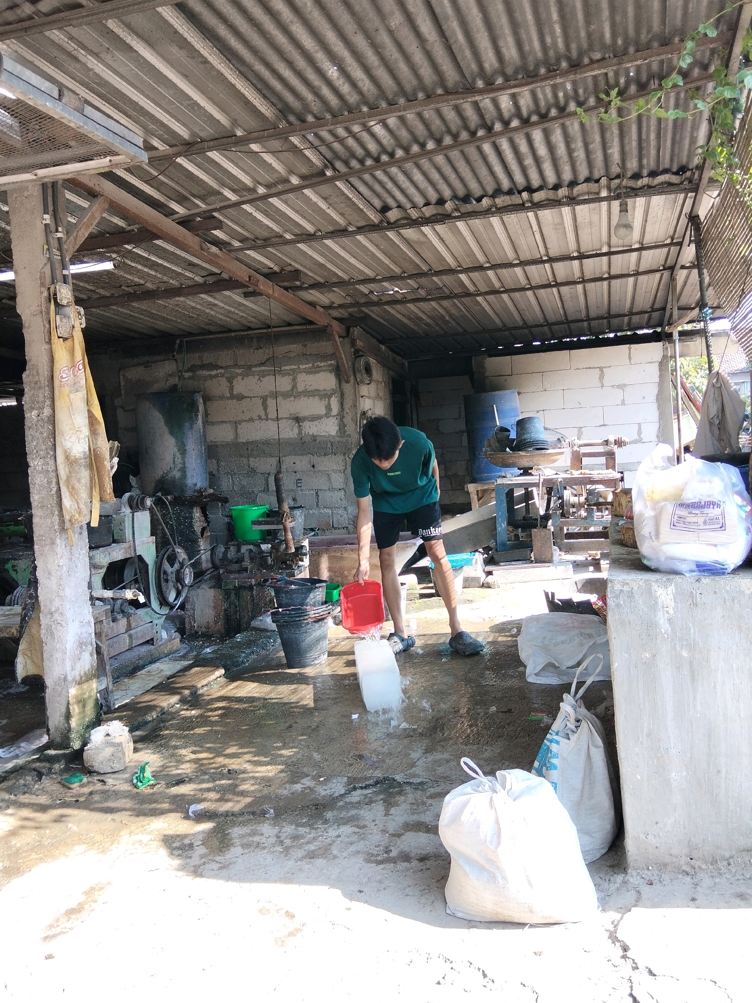In Ponorogo, Indonesia, a young male wearing a green t-shirt and black shorts is in an open-air workshop, pouring water from a red bucket onto a large block of ice on the ground. The workshop has a corrugated metal roof supported by wooden beams, and the walls are constructed of cinder blocks. Various industrial equipment, including a large machine with belts and pulleys, is visible. Several buckets, including green and black ones, are scattered around the messy, wet concrete floor. Bags of what appear to be materials are stacked near a concrete wall on the right side of the image. The environment is cluttered with tools and debris, suggesting an active workspace.