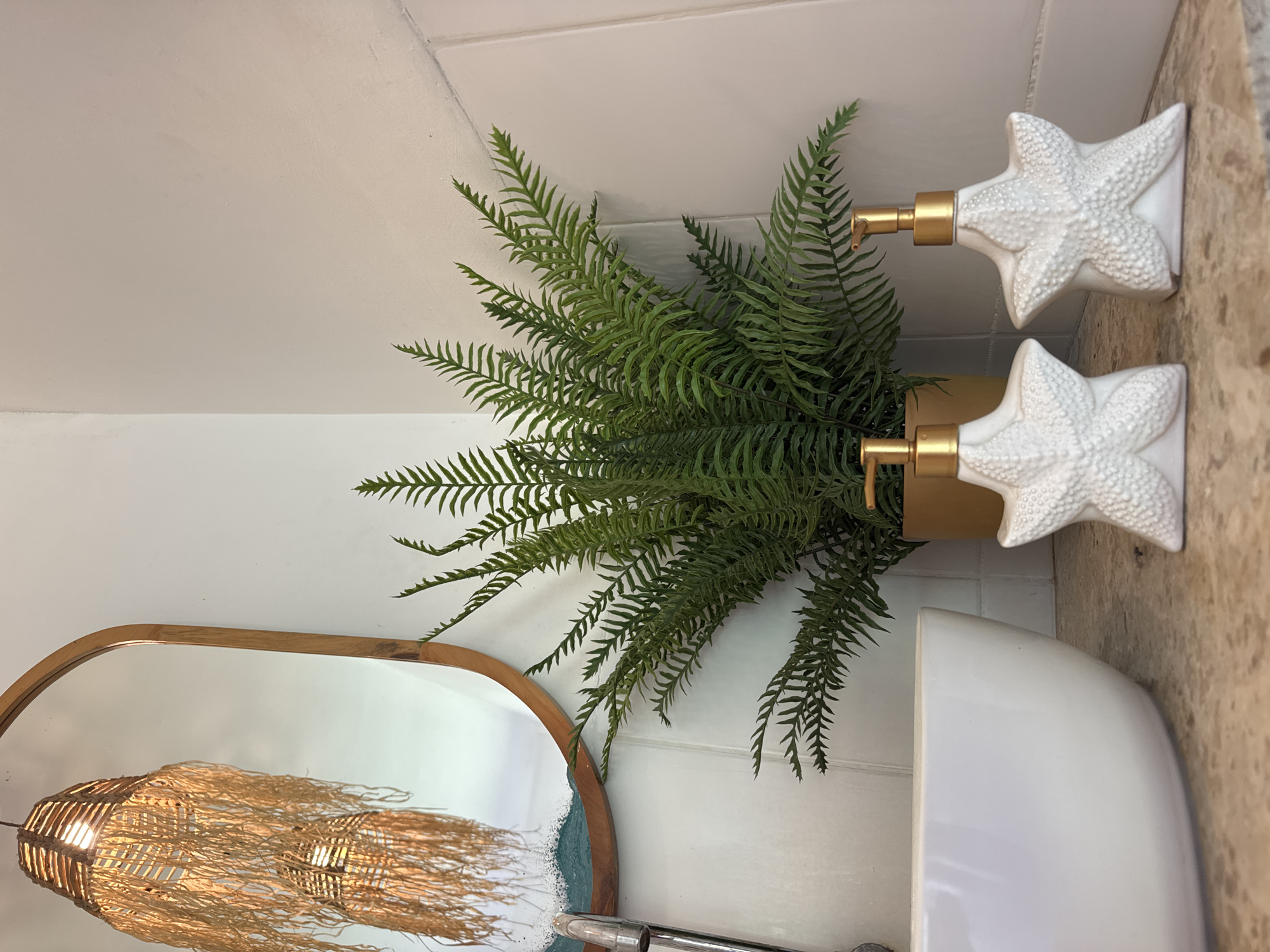 The image shows a bathroom interior, likely located in Kuta Selatan, Indonesia, given the presence of tropical-themed decor elements. The main features include a green fern plant in a metallic gold pot, positioned between two white soap dispensers shaped like starfish with gold-colored pumps. To the left, a partially visible wooden-framed mirror reflects a woven light fixture, suggesting a bohemian or coastal aesthetic. The background reveals white tiled walls and a countertop with a light brown, possibly marble-like, surface. A section of a white sink is also visible.