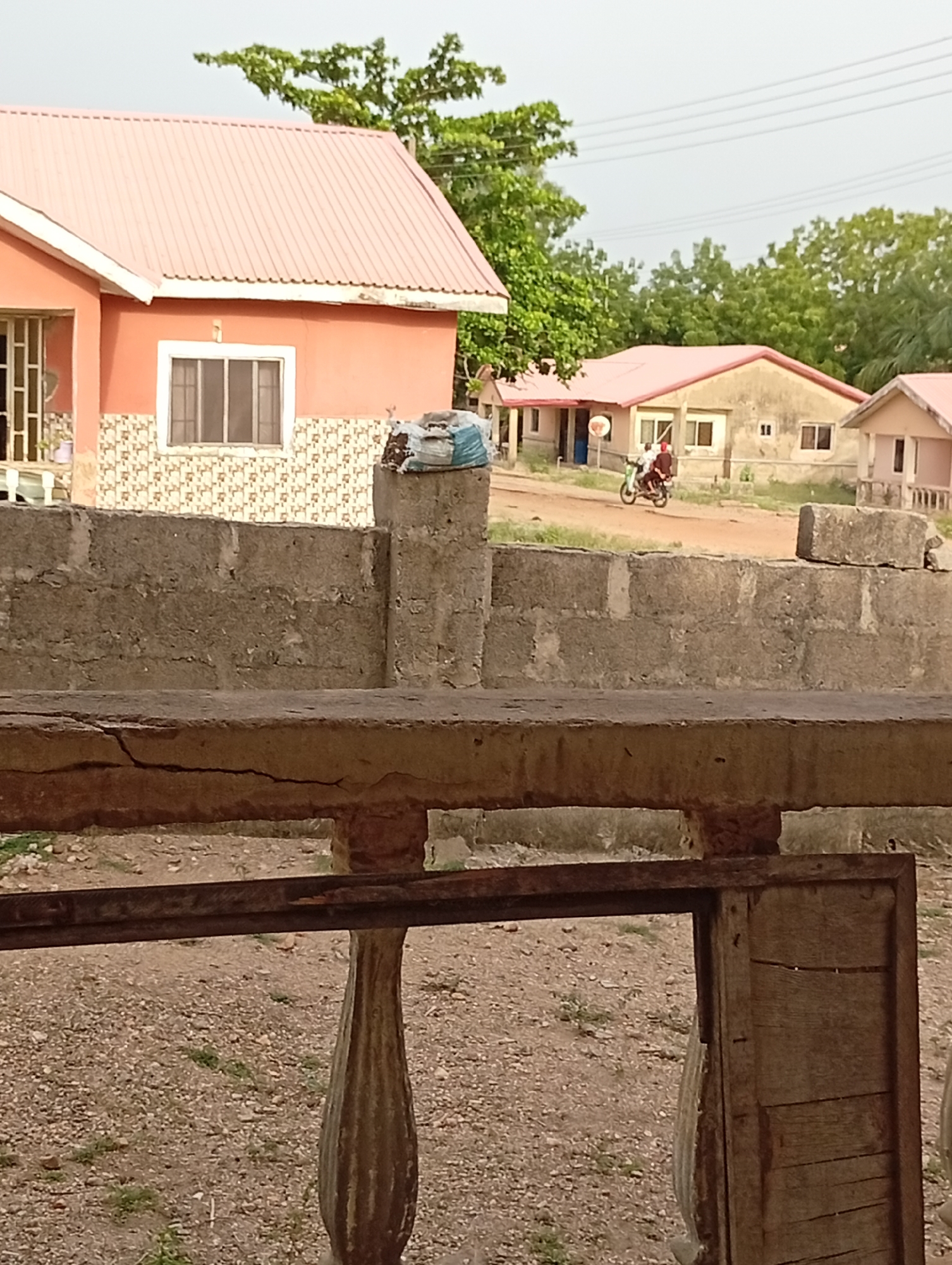 The image depicts a residential street scene in the city of Unknown, Unknown. In the foreground, a weathered wooden railing with a rusted metal frame provides a partial view. Behind the railing, a concrete wall with a stacked-stone pattern runs across the midground. On top of one of the wall's pillars sits a blue and white bag. Beyond the wall, a street scene is visible, featuring several peach-colored, single-story houses with pink metal roofs. A motorbike with two occupants is moving along the unpaved road. Trees and vegetation are interspersed throughout the scene. Overhead, utility wires stretch across the sky.