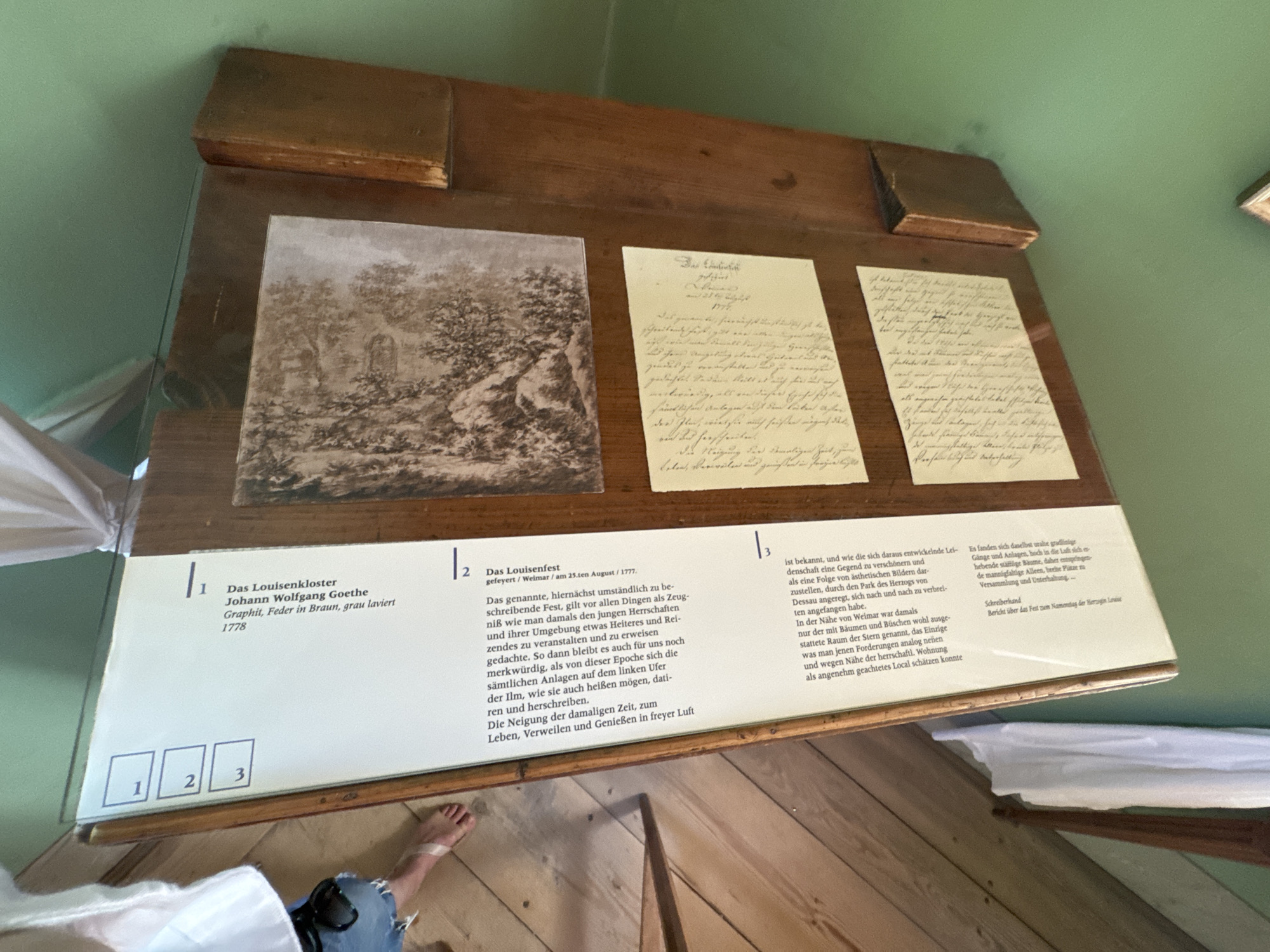 This image shows a display case within what appears to be a museum in Weimar, Germany, specifically referencing the "Das Lousienkloster." The display case contains framed text and images. On the left side of the display, there are three numbered boxes: 1, 2, and 3. The main content of the display includes a sepia-toned drawing or print of a landscape with trees and architectural elements, likely depicting the Lousienkloster. Below this image, there are two handwritten documents presented on cream-colored paper. The text on the display case is in German, with a title "Das Lousienkloster" and attributed to "Johann Wolfgang Goethe," with the medium and year "Graphik, Feder in Braun, grau, laviert 1778" indicating it's a graphic work by Goethe from 1778. Another section of text details a "Louisenfest" held on August 23, 1777, and mentions Goethe. The environment suggests an indoor exhibition space with green walls and wooden display furniture. There is a partial view of a person's legs and feet on the left side of the image, wearing denim shorts and flip-flops.
