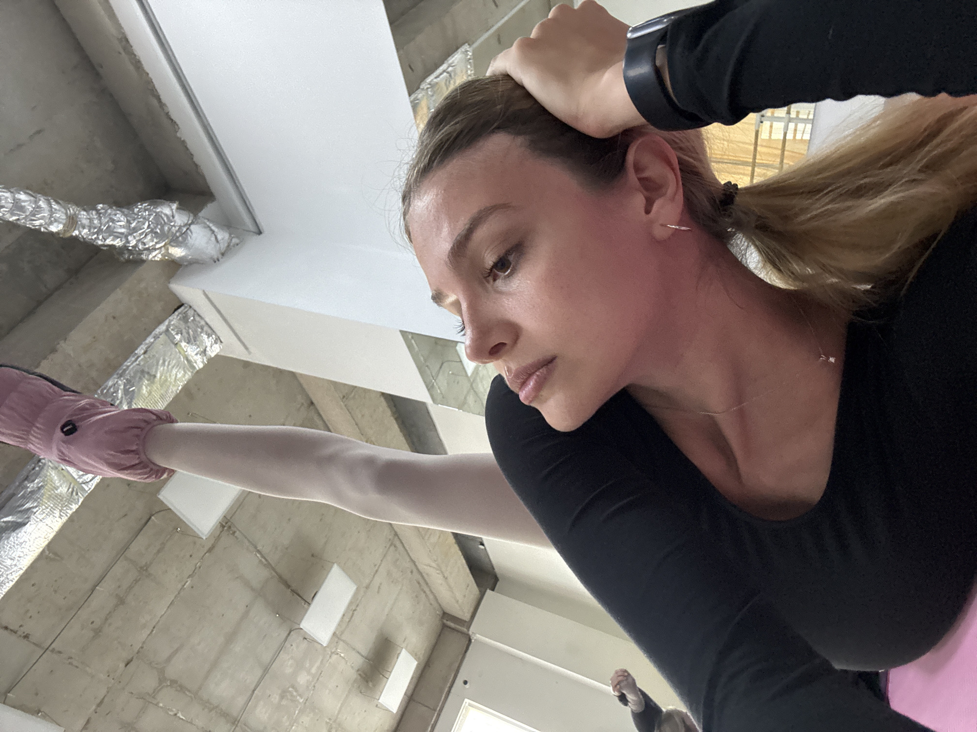 A medium-shot, eye-level image shows a fair-skinned woman performing a standing split in what appears to be an interior space. She is wearing a black long-sleeved top, and her blonde hair is pulled back into a ponytail. Her left arm is extended, holding her head, and she is wearing a dark smartwatch on her left wrist. Her left leg is extended upwards with a pink sock-like covering on her foot.
The background consists of what seems to be an unfinished room or warehouse. There are exposed concrete ceilings with metal ductwork wrapped in foil insulation. Several white rectangular light fixtures are visible on the ceiling. A partial view of a door is seen in the lower part of the image. Another individual is visible in the lower-right corner of the image, appearing blurry and further away.
Given the industrial appearance and urban setting, it is plausible the location is in the city of Tbilisi, Georgia, though this cannot be definitively determined from the image.