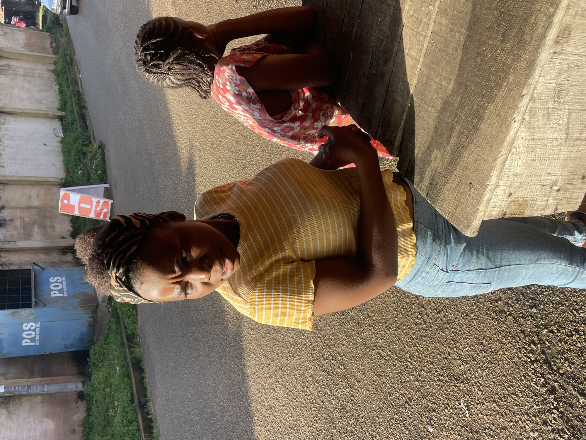 A young Black woman with her hair in a patterned head wrap and a yellow and white striped shirt is standing in front of a wooden table. She is looking away from the camera with a concerned expression. Behind her, another young Black woman with braided hair and a pink floral dress is leaning on the table, facing away from the camera. The background shows a street in what appears to be a commercial area, with a building featuring "POS" signage, indicating Point of Sale services, likely in the city of Jos, Nigeria. The ground is gravelly, and there is a road with a vehicle visible in the distance. The lighting suggests it is daytime with strong sunlight creating shadows.