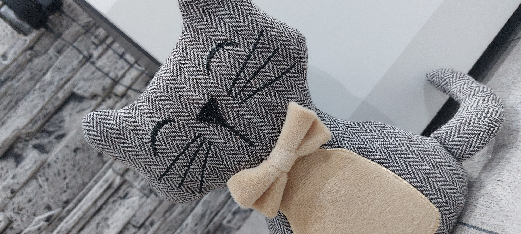 A plush cat toy lies on a surface. The toy is made of a herringbone pattern fabric in gray and white. The cat has a triangular nose and whiskers stitched in black. It also has a beige bow tie. The background is a combination of a gray brick pattern on the left and a white surface on the right. The location is unknown and cannot be determined from the image. The city of Yagodovo, Bulgaria is also unknown from the image.