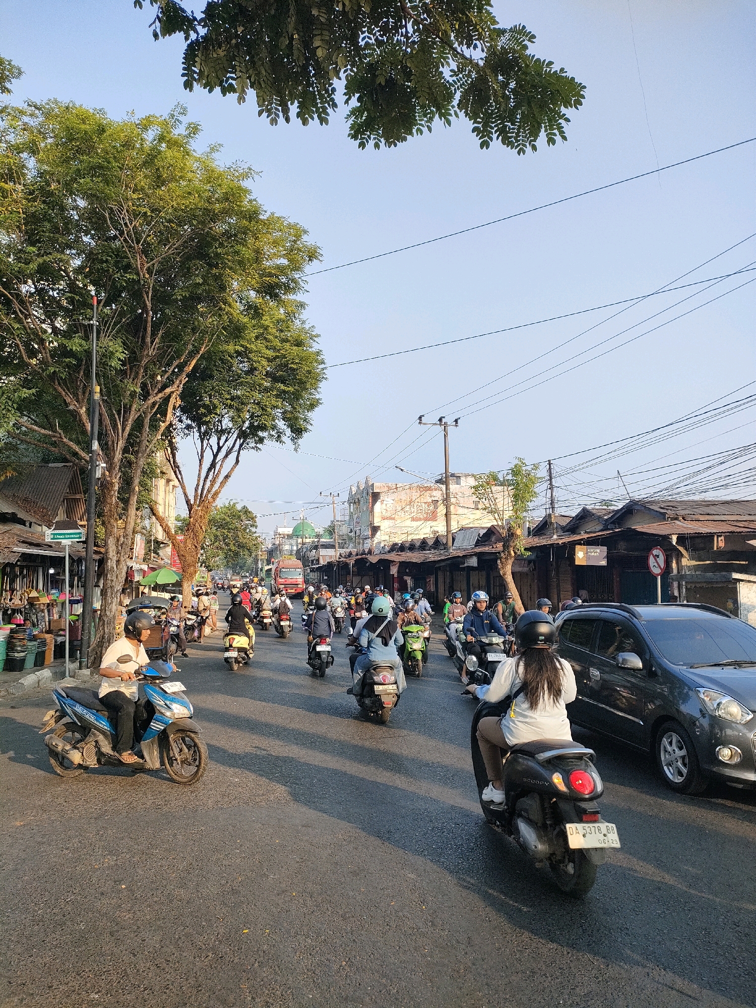 This is a street scene in Banjarmasin, Indonesia, likely taken during the day due to the bright sunlight casting shadows. The image depicts a busy urban environment with a considerable amount of motorcycle traffic. Many individuals are riding scooters or motorcycles, most wearing helmets. On the left side of the street, there are market stalls or shops with awnings and goods displayed, suggesting a commercial area. A prominent green tree with dense foliage is on the left, casting shadows on the road. Further down the street, buildings with traditional architecture and signs are visible, including what appears to be a mosque with a green dome. Overhead, numerous electrical wires crisscross the sky. A dark-colored car is also present on the right side of the road, adding to the variety of vehicles. The road surface is asphalt, and the overall atmosphere appears to be one of daily life and commerce in the city.
