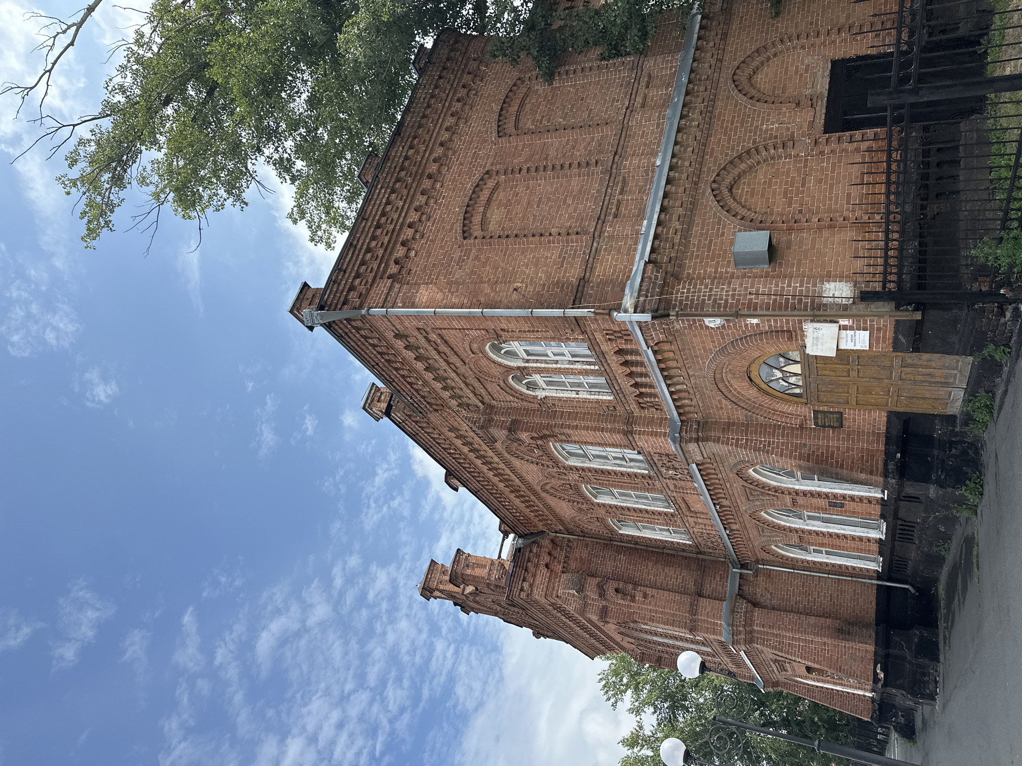 An exterior low-angle shot shows a large, ornate brick building against a bright blue sky with scattered clouds. The building is constructed of red brick with decorative elements, including arched windows and intricate brickwork along the roofline and around the window frames. A tall, pointed architectural feature adorns the left side of the building. To the right, a large tree with green foliage partially obscures the building. In the foreground, a paved walkway runs alongside the building, with a black metal fence bordering it. A lamppost with two globe-shaped lights is visible on the left. The building appears to be in a state of disrepair in some areas, with visible water damage at the base and faded paint. A wooden door is set into an arched entryway on the right side of the building, with a sign attached to the wall next to it.