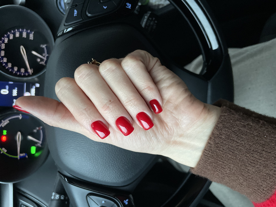 A hand with freshly manicured, square-shaped red nails rests on a black steering wheel. A gold ring adorns the ring finger. The dashboard, including a speedometer and digital display, is visible in the background. A brown sweater cuff frames the right side of the image. The car's interior is dimly lit.
