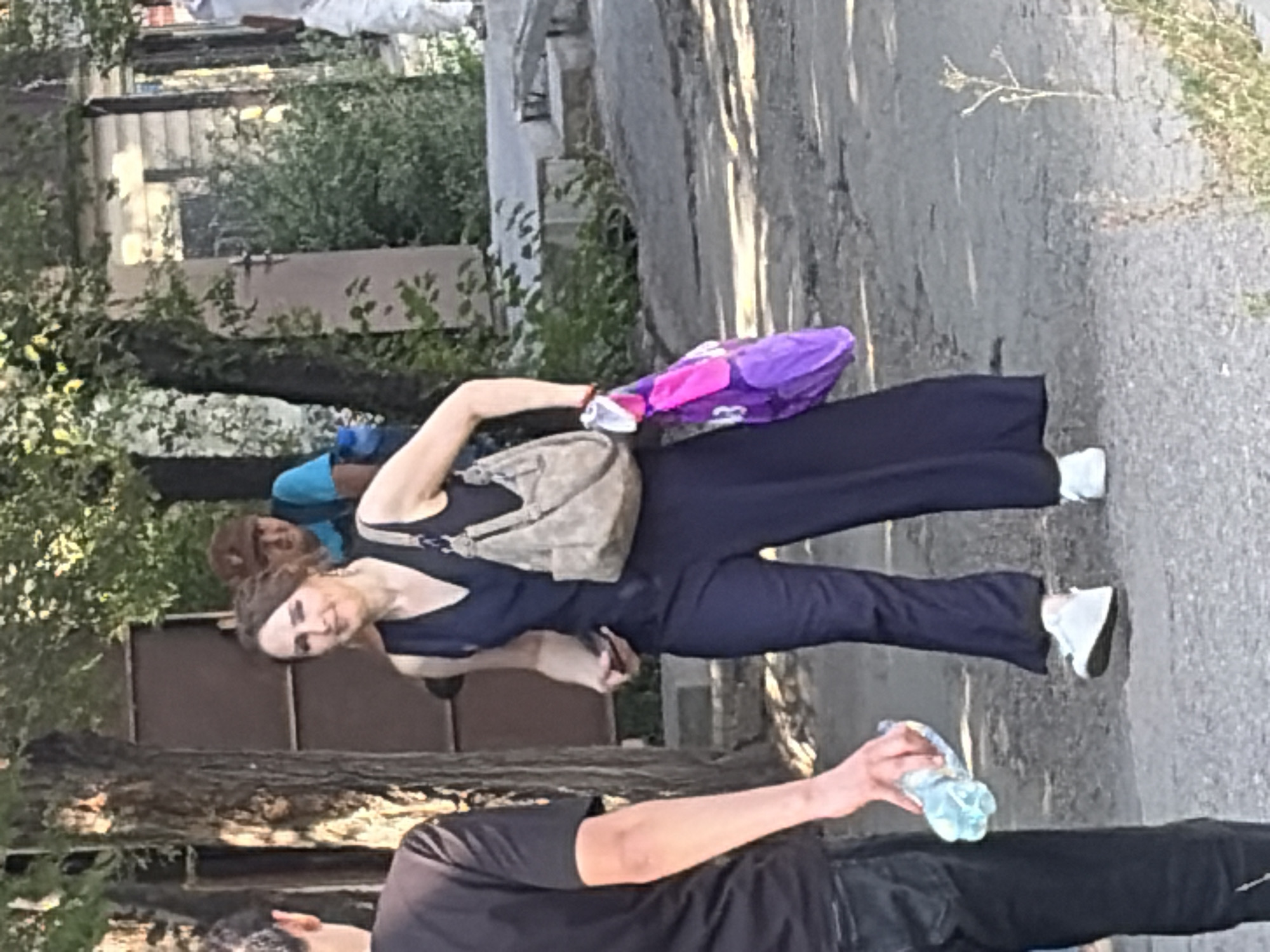 In the city of Unknown, Unknown, an outdoor scene unfolds on a paved surface with trees and buildings in the background. A woman, seen from the knees down, is wearing a dark blue jumpsuit and white sneakers. She is holding a purple bag with a pink strap in her left hand and a light brown purse on her right shoulder. To her left, a man, also seen from the knees down, is wearing dark pants and holding a clear plastic bottle of water in his right hand. Behind the woman, two other individuals are visible: one wearing a blue t-shirt and a brown hat, and another person further back, whose details are obscured. The environment appears to be a street or pathway with uneven ground and scattered vegetation. The lighting suggests it is daytime, with sunlight casting shadows.