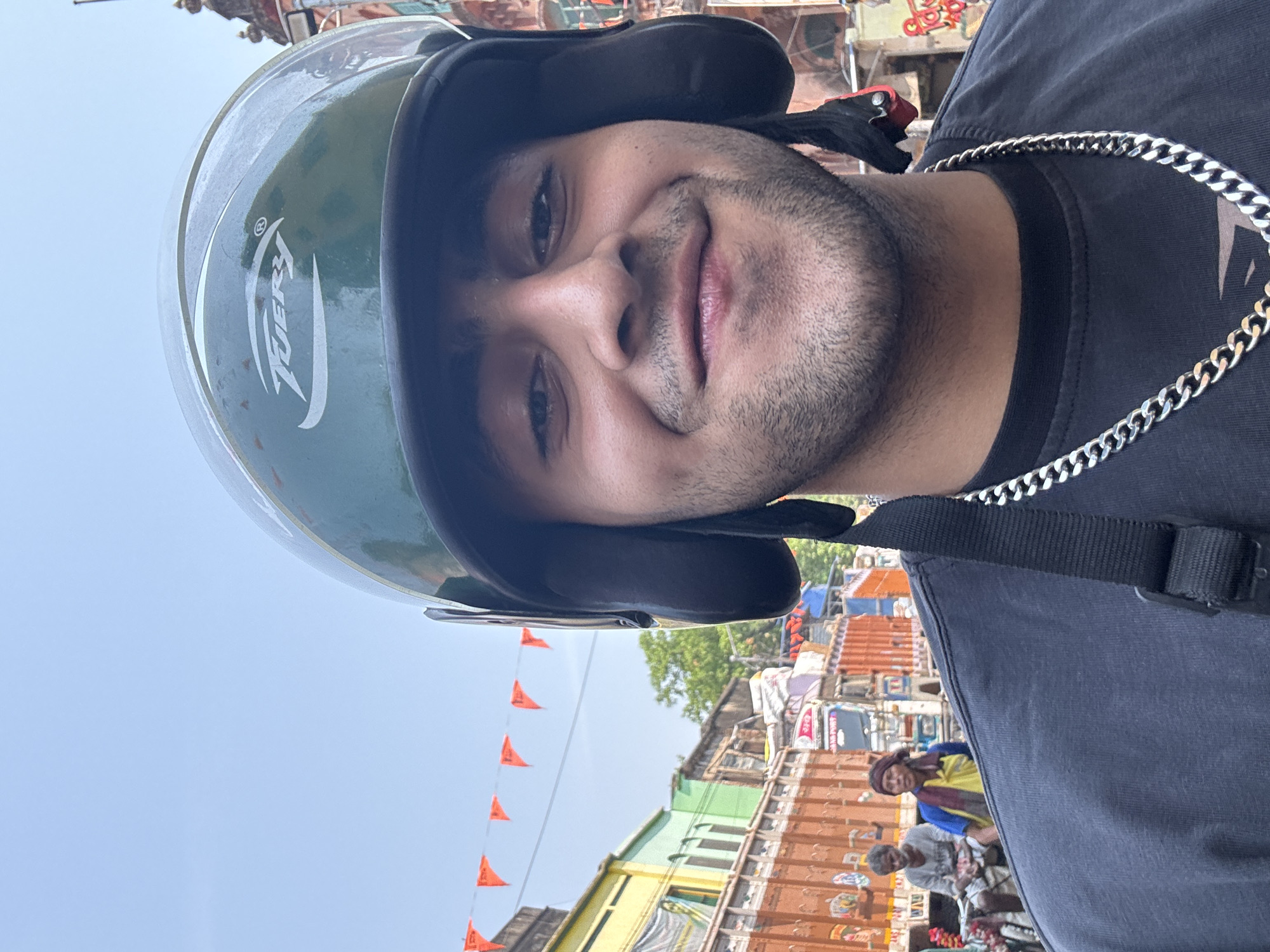 A man wearing a green helmet and silver chain is visible in the foreground. He is also wearing a black t-shirt and has a black strap running across his body. He is smiling. In the background, there is a cityscape with buildings of varying heights and colors. Red flags are strung across the sky. People and vehicles are present on the street. The scene appears to be in Kolkata, India, based on the architecture and overall urban environment.