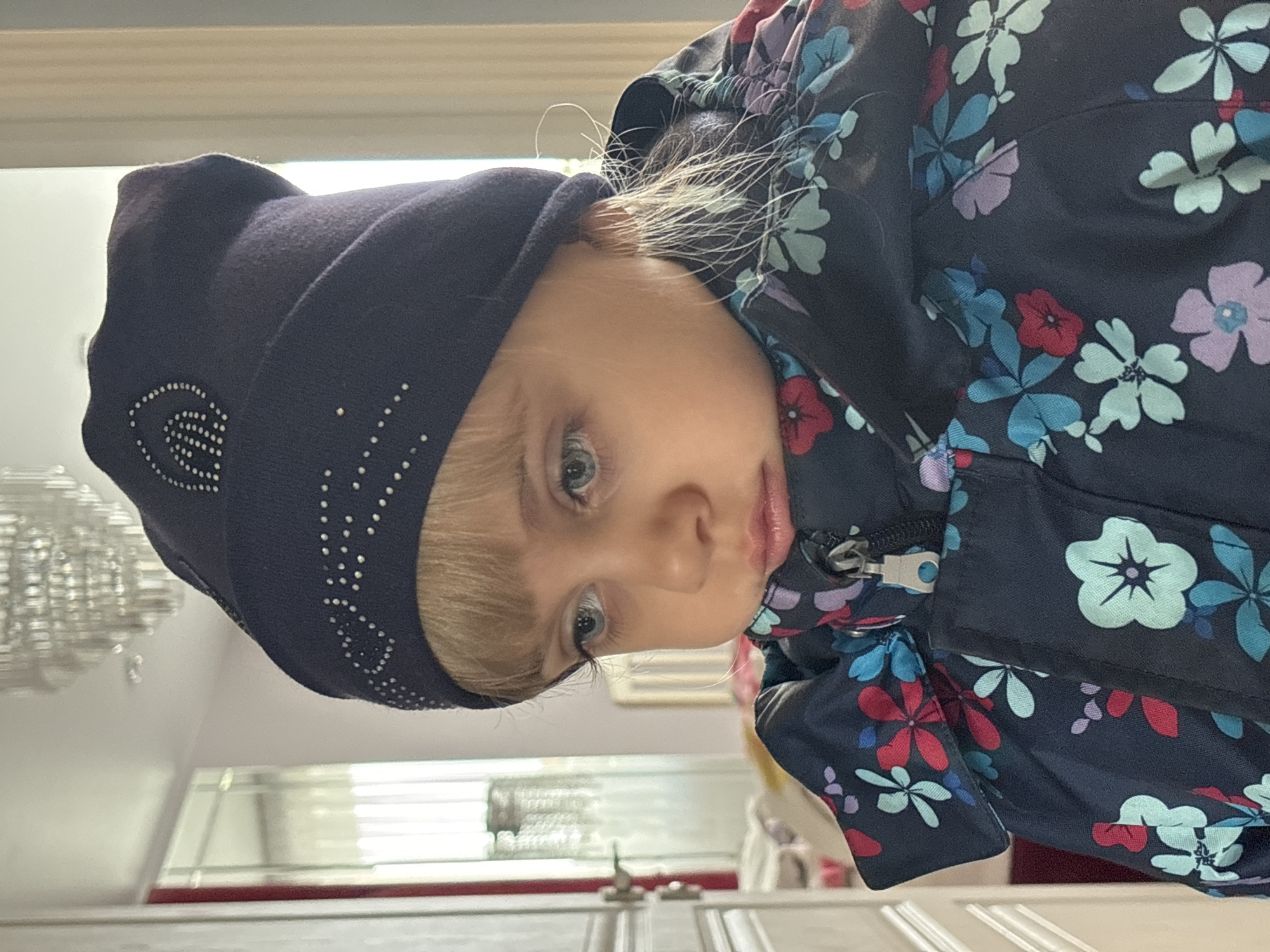 A close-up shot of a young child is captured, showing the subject from the chest up. The child is angled slightly to the right, wearing a dark blue beanie with decorative bead accents and a floral-patterned jacket, zipped near the neck. The child has fair skin, light blonde hair, and blue eyes, gazing towards the viewer. The background is somewhat blurred, hinting at an interior setting with a chandelier and white cabinets with silver hardware. The location is Unknown, Unknown.