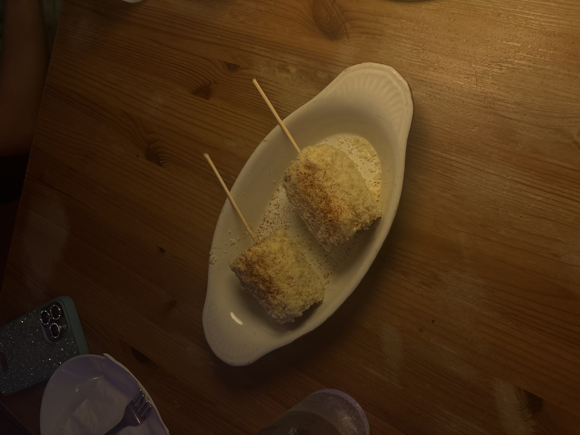 Two pieces of corn on the cob, each with a skewer inserted, are presented on a white oval dish. The corn appears to be coated in a light-colored crumb or cheese mixture and sprinkled with red seasoning. The dish is placed on a wooden table. To the left of the dish is a partially visible glass of water and a white plate with a fork. In the upper left corner of the image is a glittery teal phone case. The scene is dimly lit, suggesting an indoor setting, possibly a restaurant. The image does not provide any specific information to determine the location within San Juan, United States of America.