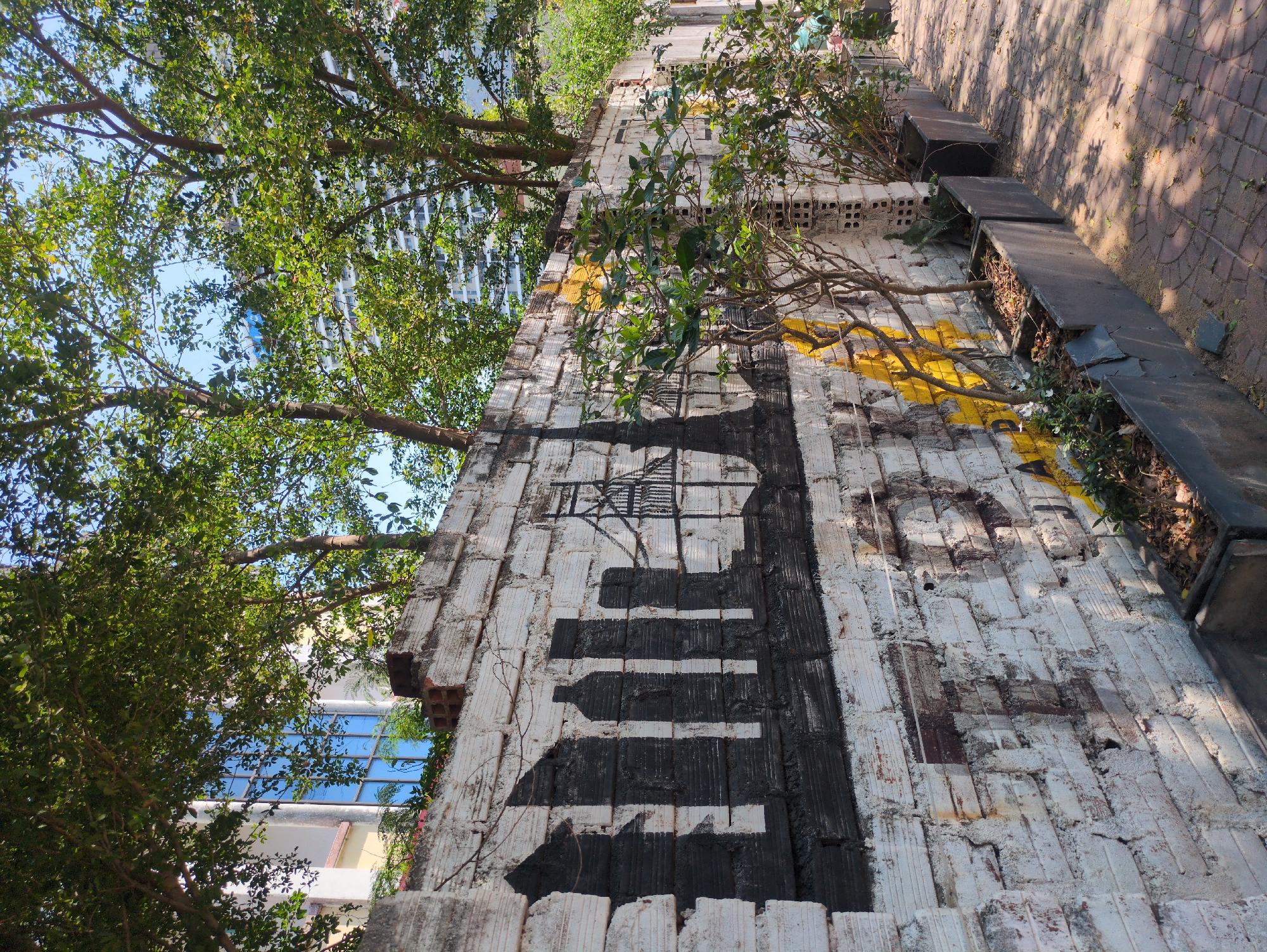 The image shows a long, low brick wall in Quy Nhon Ward, Vietnam, with a piano keyboard painted on it in black and white. The wall appears weathered and aged, with some areas of crumbling brick. Trees are growing alongside the wall, partially obscuring the keyboard design and casting shadows on the wall. The keyboard is slightly distorted, painted on the uneven surface of the brick. The black keys are solid rectangles, while the white keys are represented by the gaps between them. The tops of the brick wall contain soil and some vegetation growing along the side of it. Some rectangular metal planters are placed along the end of the wall. The area adjacent to the wall is paved with red bricks. A tall building is visible in the background through the foliage.