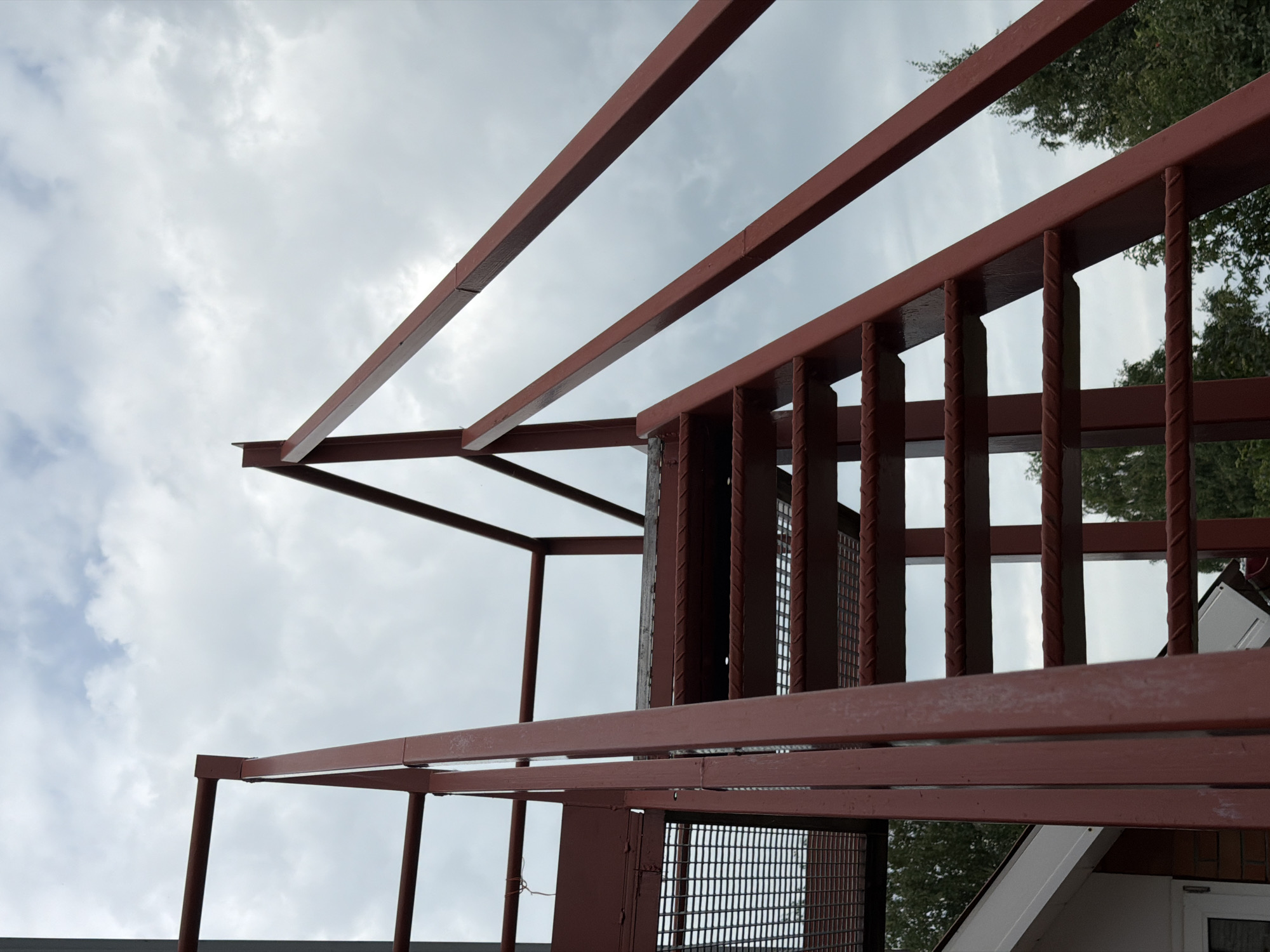 A low-angle, close-up shot captures a section of a red metal staircase against a cloudy sky. The staircase, constructed from sturdy metal beams and featuring a grated walkway, ascends diagonally from the left side of the frame. Several steps are visible, their surfaces reinforced with textured bars. A metal railing runs along the right side of the stairs. In the background, the top of a white-capped building is visible to the left, with a section of brick wall and a closed window peeking out. Trees with green foliage are seen behind and below the staircase, suggesting an outdoor setting. The overall impression is one of utilitarian construction against a natural, albeit overcast, backdrop. The specific location in Tenishevo, Russia, is not visually discernible from the image itself.
