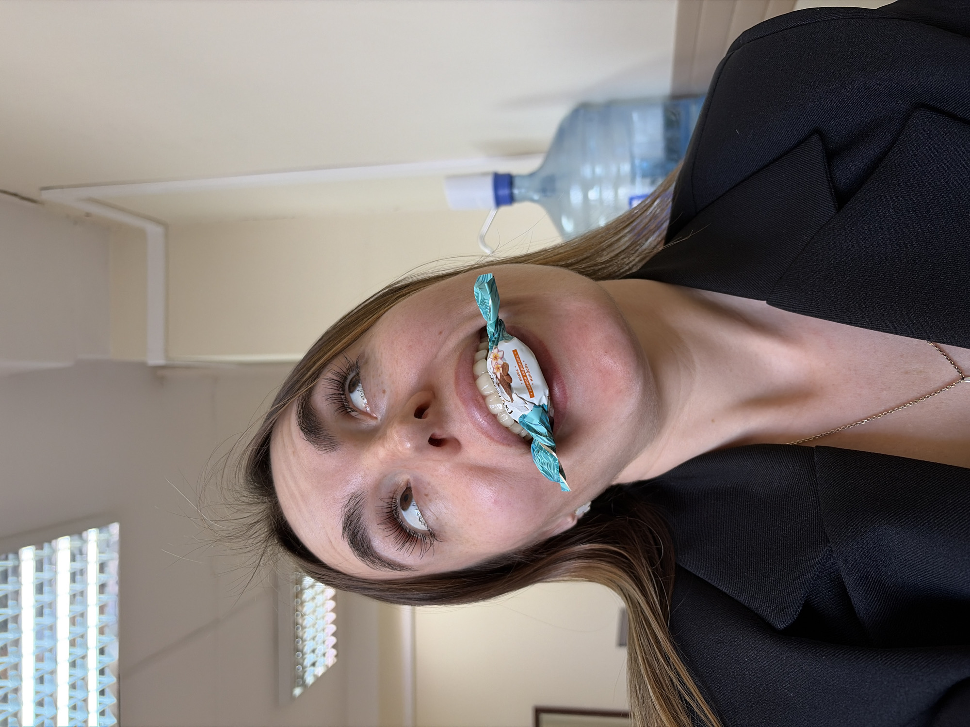 A woman is depicted in a photograph, holding a wrapped candy in her mouth. The woman has brown hair and is wearing a black blazer with a gold necklace. A large bottle of water is visible in the background. The indoor environment appears to be an office setting, based on the ceiling structure and lighting. The city of Izhevsk, Russia, is not evident in the image.