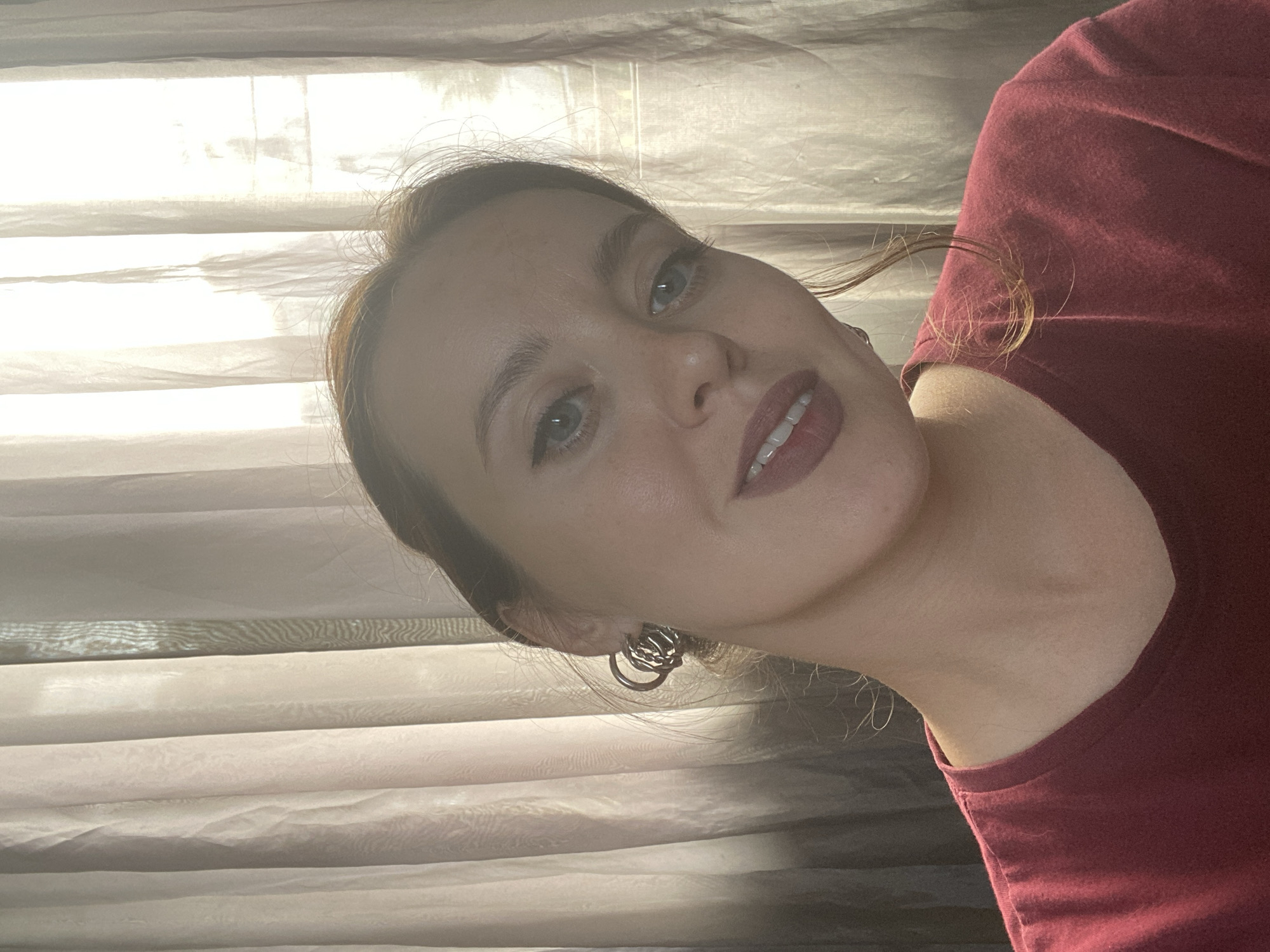 A young woman with light skin and auburn hair tied back is looking at the camera. She is wearing a maroon t-shirt and large, circular, silver earrings. Her lips are painted a deep pink. She is positioned in front of sheer, cream-colored curtains. The light is coming from behind the curtains, creating a bright, almost blown-out effect in the background and casting shadows. The setting appears to be an indoor space, possibly a home or hotel room, with the curtains suggesting a window is present. There is no clear indication of the location being in the city of Batumi, Georgia.