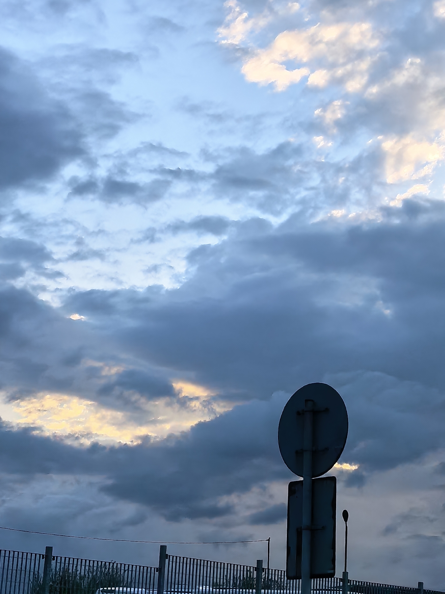 The image captures a dramatic sky filled with various shades of blue and gray clouds, suggesting an overcast day with moments of light. Patches of warm, golden-yellow light peek through openings in the darker cloud formations, indicating either dawn or dusk.
In the lower right foreground, the back of a dark, circular road sign is prominently visible, attached to a vertical pole. Below this, a section of a dark metal fence or railing extends horizontally across the lower part of the frame. Behind the fence and to the right of the sign, a thin, dark pole, possibly a lamp post, stands.
There is no discernible activity taking place; the scene is static, focusing on the atmospheric conditions and foreground objects. No visible text is present on the road sign or elsewhere in the image. The overall impression is one of a transitional time of day, possibly from a road or elevated structure.
