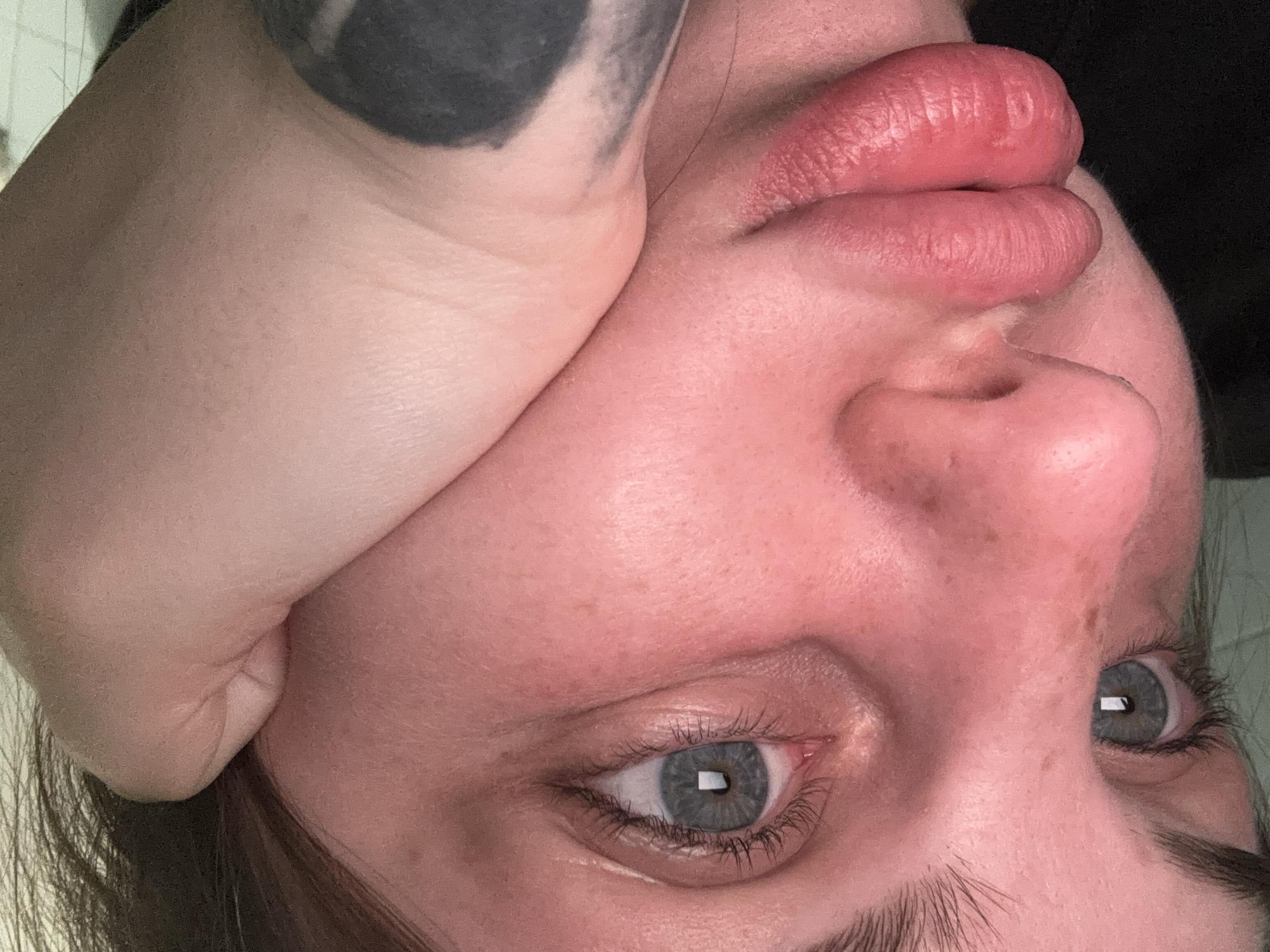 A close-up, upside-down perspective of a person's face is presented. The skin appears flushed or red. The person has blue eyes and light eyebrows. Their lips are full and reddish. Part of a hand with a dark tattoo is visible near the top left of the frame. The background is dark. The location cannot be determined from the image.