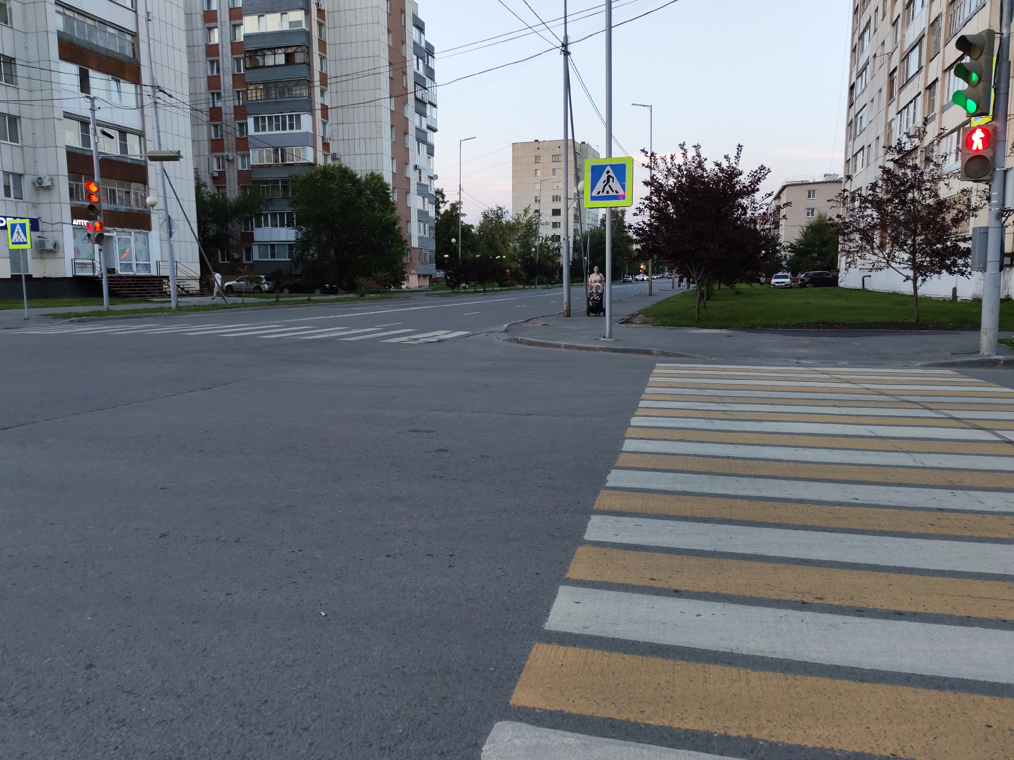 The image depicts a street scene at an intersection in a city. In the foreground, a pedestrian crossing is visible, with alternating yellow and white stripes. To the left of the crossing, a traffic light displays a red signal for pedestrians, indicated by a red walking figure, and a red light for vehicles. Further to the left, a blue and yellow pedestrian crossing sign is mounted on a pole.
Dominating the left side of the image is a multi-story apartment building with a light-colored facade and visible balconies. Across the street, on the right side, another similar apartment building stands. Between these buildings, trees with dark foliage are planted.
In the middle ground, the street curves, and a few vehicles are parked or driving. A woman pushing a stroller is visible on the right side of the street, near a pedestrian crossing sign. The sky is overcast, suggesting dusk or an early evening hour. Power lines and streetlights are present throughout the scene.