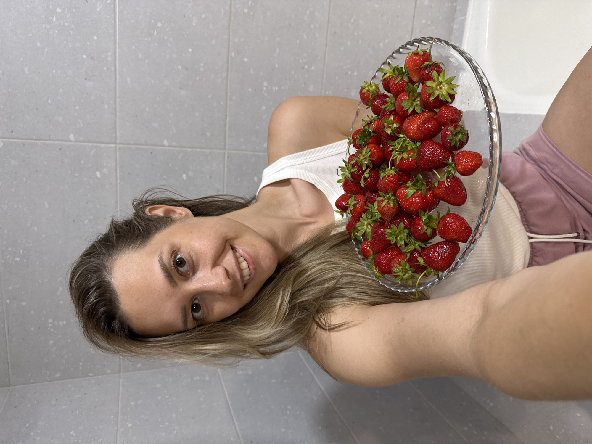 A young woman with blonde hair is holding a clear glass plate filled with strawberries. She is wearing a white tank top and pink shorts. The background appears to be a bathroom with grey tiled walls and a white bathtub. The woman is smiling at the camera, taking a selfie. The scene is indoors, likely in a residential setting, given the bathroom environment. The specific location within the city of Nebug, Russia cannot be determined from the provided image alone.